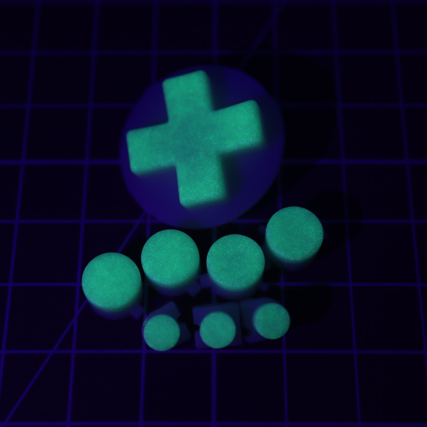 The Analogue Pocket - Custom Button - GITD Green, including glow-in-the-dark cylindrical buttons and a plus-shaped piece created with handmade molds, is carefully placed on a grid-lined surface.