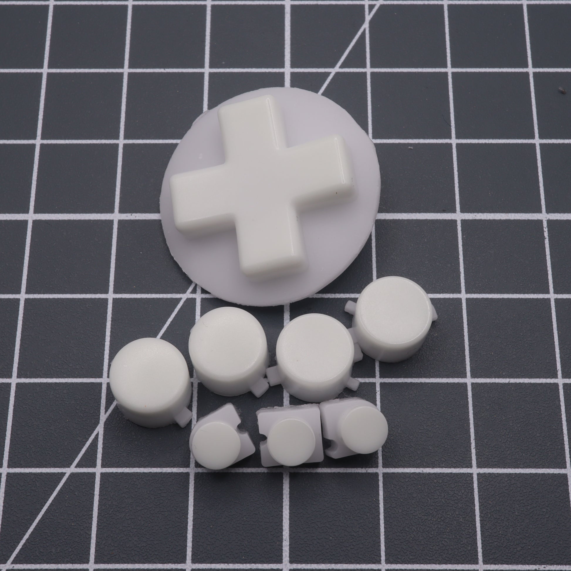 Glow-in-the-dark blue custom buttons on a black grid mat, with one large cross-shaped piece and smaller circular ones, similar to Analogue Pocket buttons. Likely crafted from handmade molds for a unique touch to your gaming controller setup.