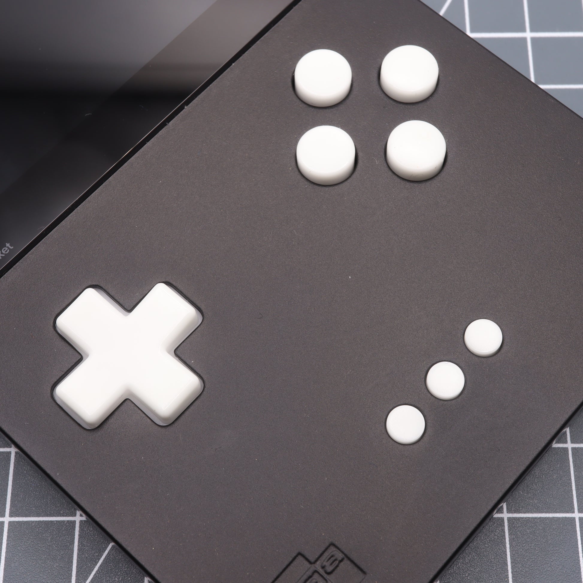 Close-up of the Analogue Pocket, a black handheld gaming console featuring GITD Blue custom-cast buttons, including a D-pad and four circular buttons, set against a grid-patterned surface.