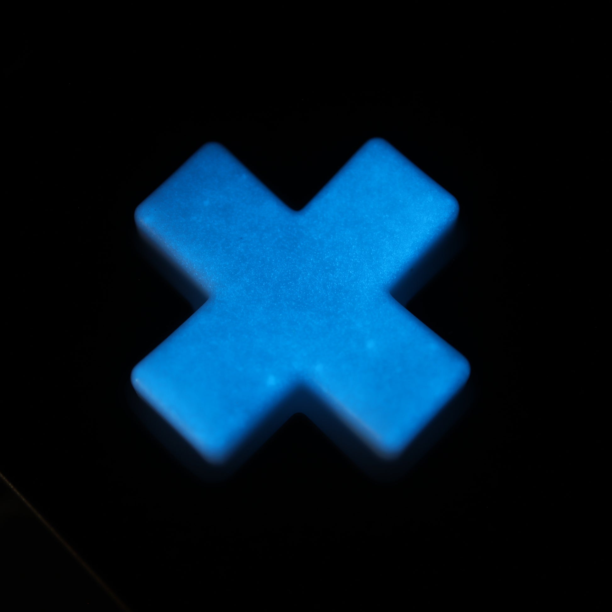 A blue glowing cross-shaped object on a dark background resembles the Analogue Pocket - Custom Button - GITD Blue, crafted using handmade molds.