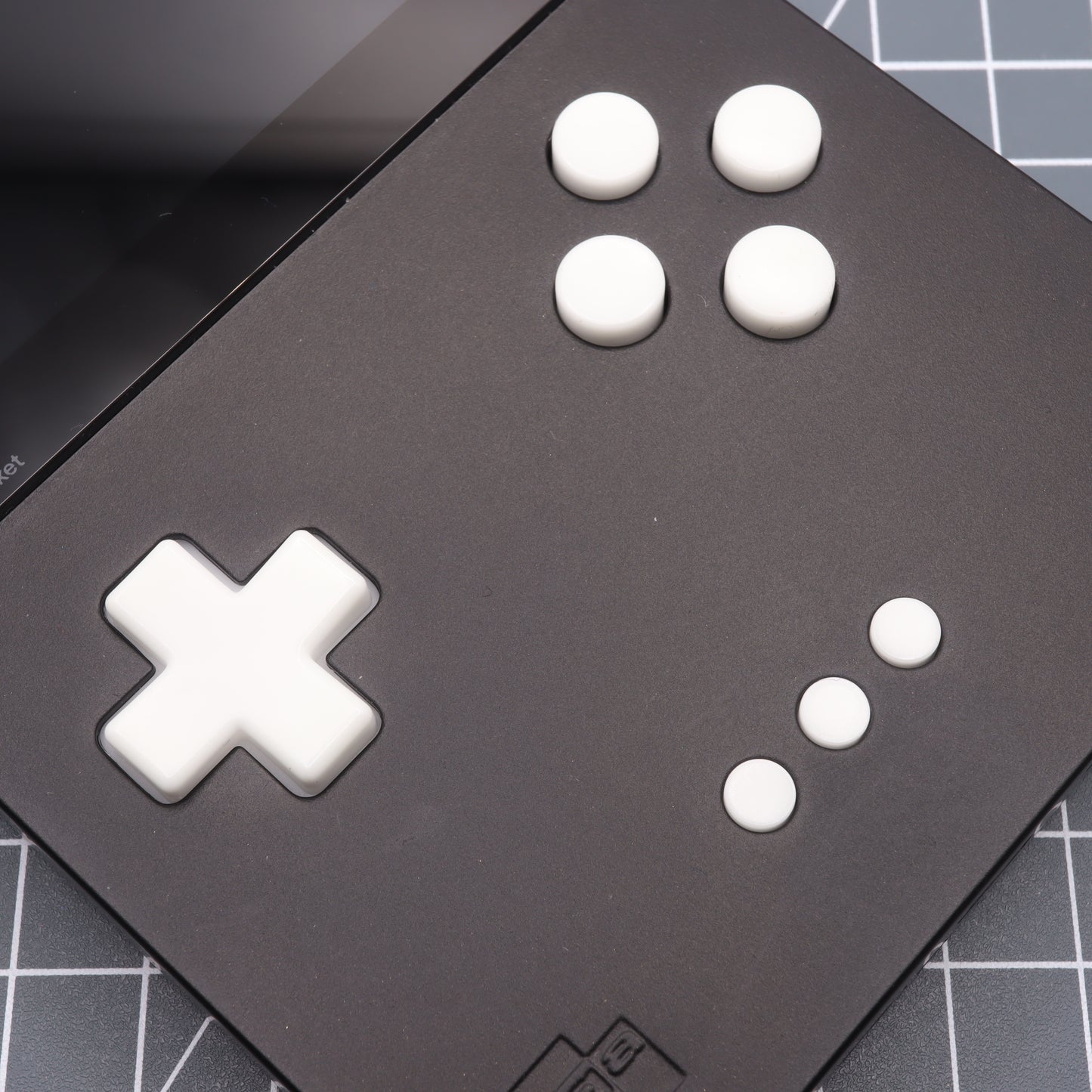 Close-up of the Analogue Pocket - Custom Button - GITD Aqua, a handheld gaming console with a black casing, featuring a white directional pad and five custom-cast buttons on a grid-patterned surface.