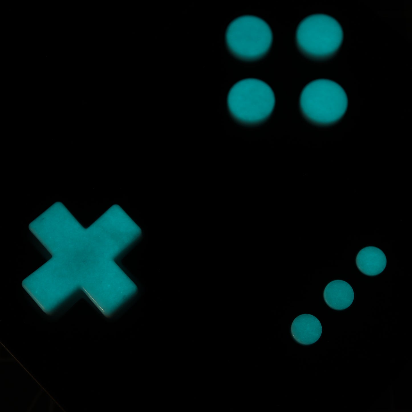 A glowing turquoise cross and six circles crafted from Analogue Pocket's custom-cast GITD Aqua buttons are arranged on a black surface, resembling parts of a classic gaming controller.