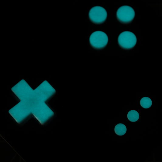 A glowing turquoise cross and six circles crafted from Analogue Pocket's custom-cast GITD Aqua buttons are arranged on a black surface, resembling parts of a classic gaming controller.