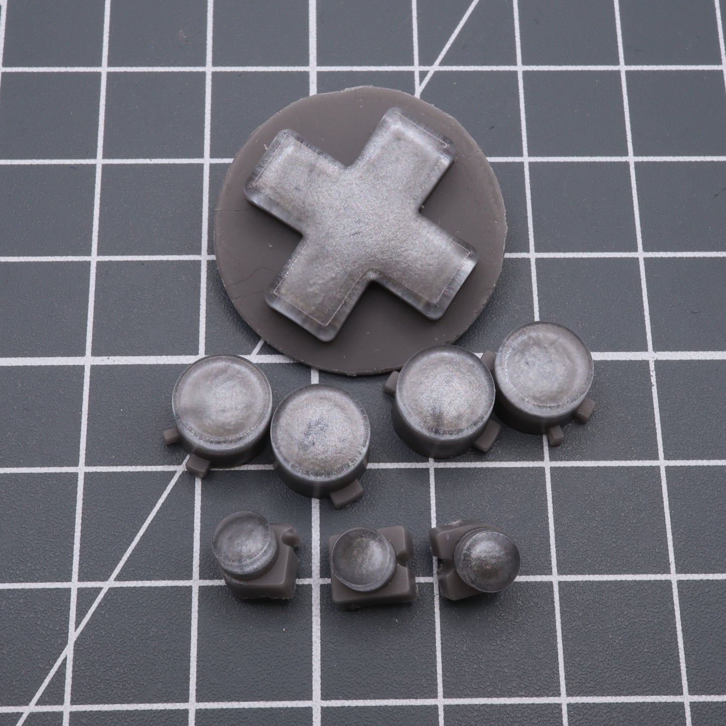 A collection of gray plastic model parts on a grid-patterned surface features a large cross-shaped piece and seven circular components, reminiscent of Analogue Pocket's Custom Button in Metallic Silver made with handmade molds.