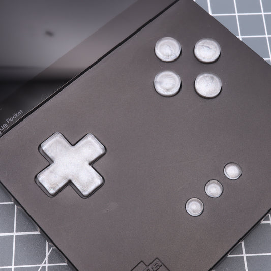 Close-up of a handheld gaming device featuring Analogue Pocket's custom metallic silver buttons and a D-pad, resting on a grid-patterned surface.