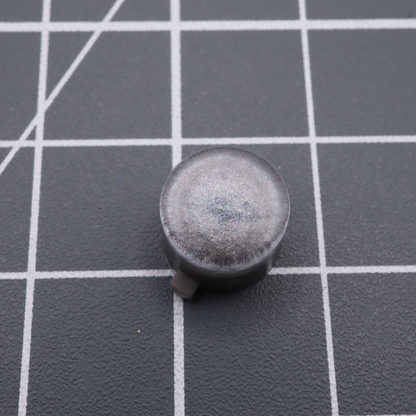 An Analogue Pocket - Custom Button in Metallic Silver, small and round like a custom-cast button, rests on a grid-patterned surface.