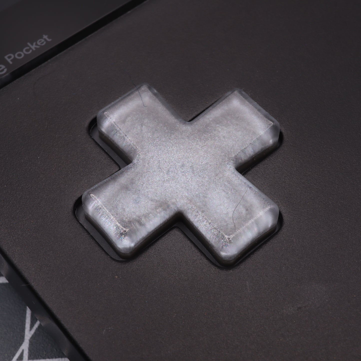 Close-up of a metallic silver cross-shaped button from Analogue Pocket, custom-cast with impeccable detail, sitting on a dark surface.