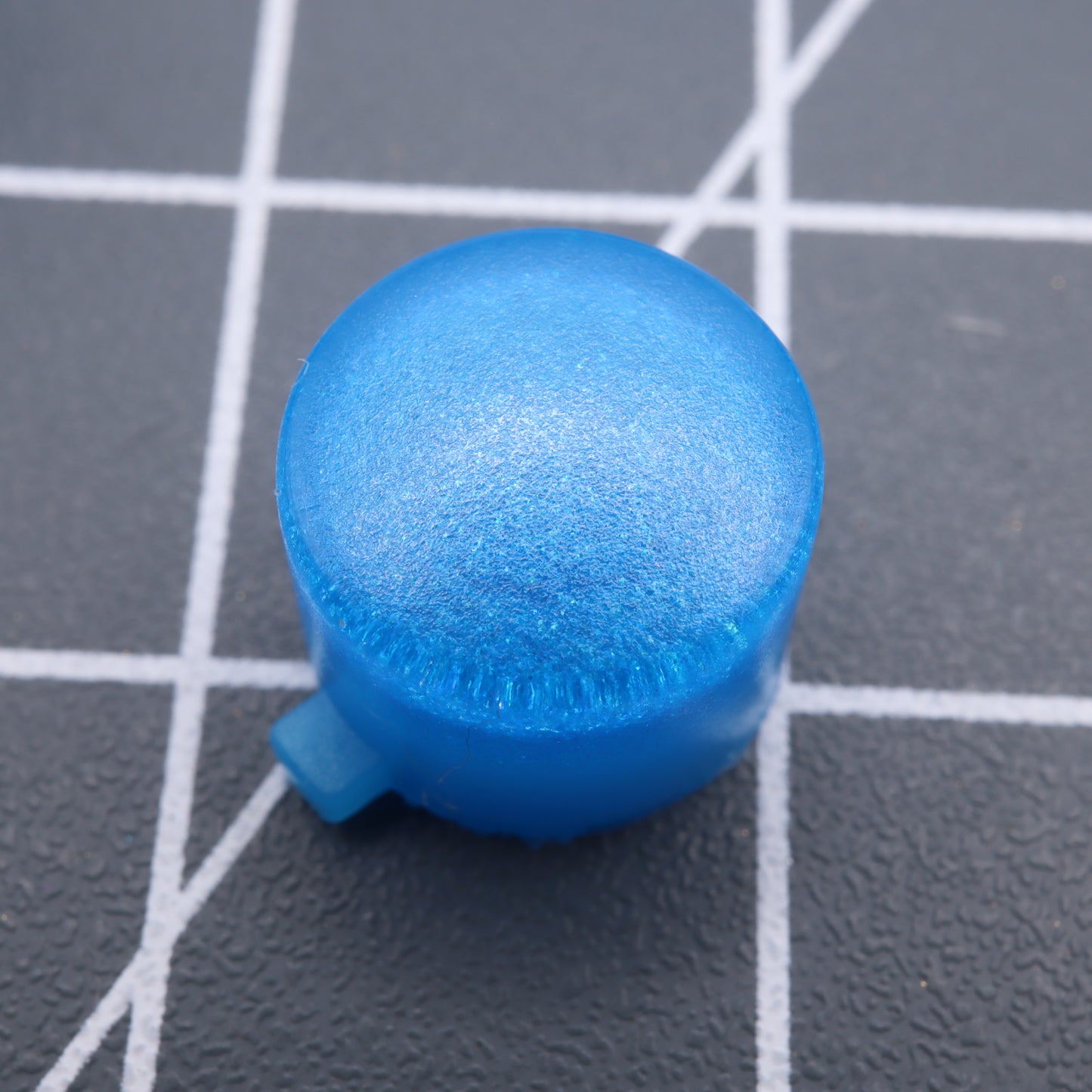 Close-up of a blue plastic cap on a textured gray surface with white grid lines, similar to Analogue Pocket's Blueberry Candy custom-cast buttons.