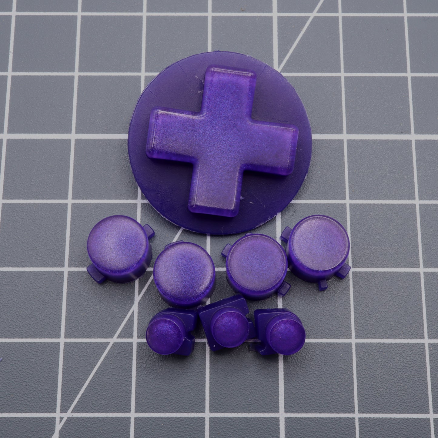 The Analogue Pocket - Custom Button - Grape Candy features custom-cast purple buttons and a D-pad resting elegantly on a grid-patterned surface, exemplifying the craftsmanship of handmade molds.