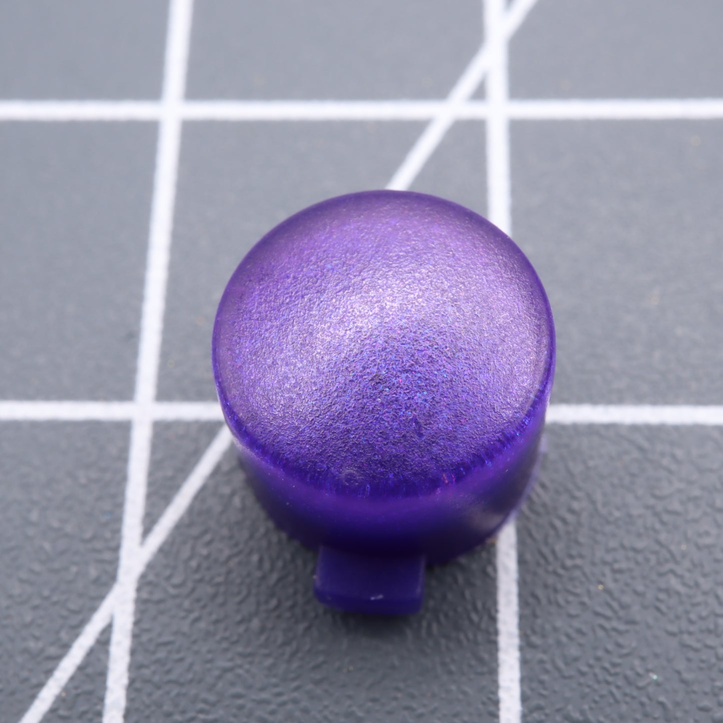 A Grape Candy Analogue Pocket custom button, a purple cylinder with a rounded top, rests on a gray grid-patterned surface, reminiscent of handmade molded designs.