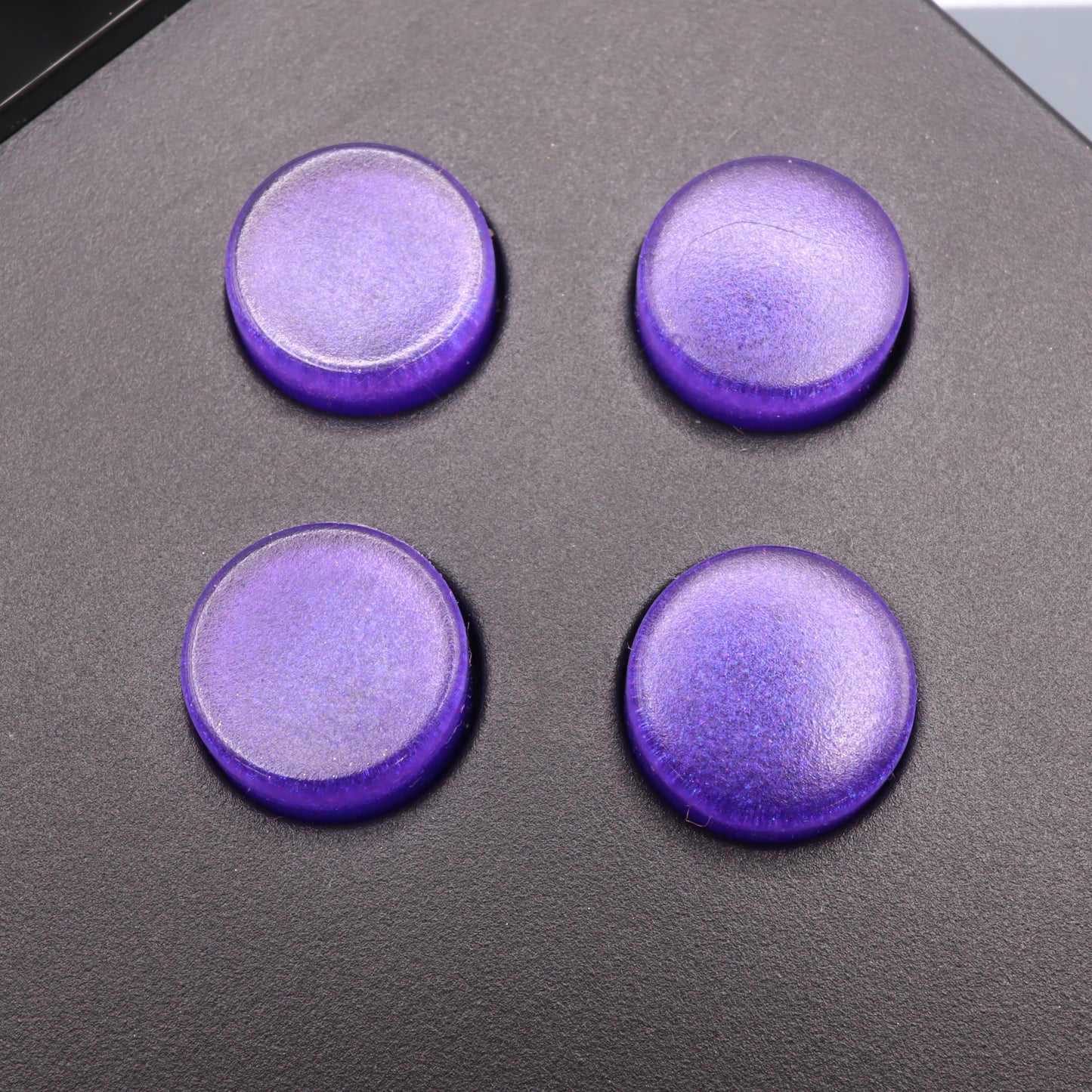 On a sleek black surface sit four Analogue Pocket Custom Buttons in Grape Candy, circular and perfectly crafted.