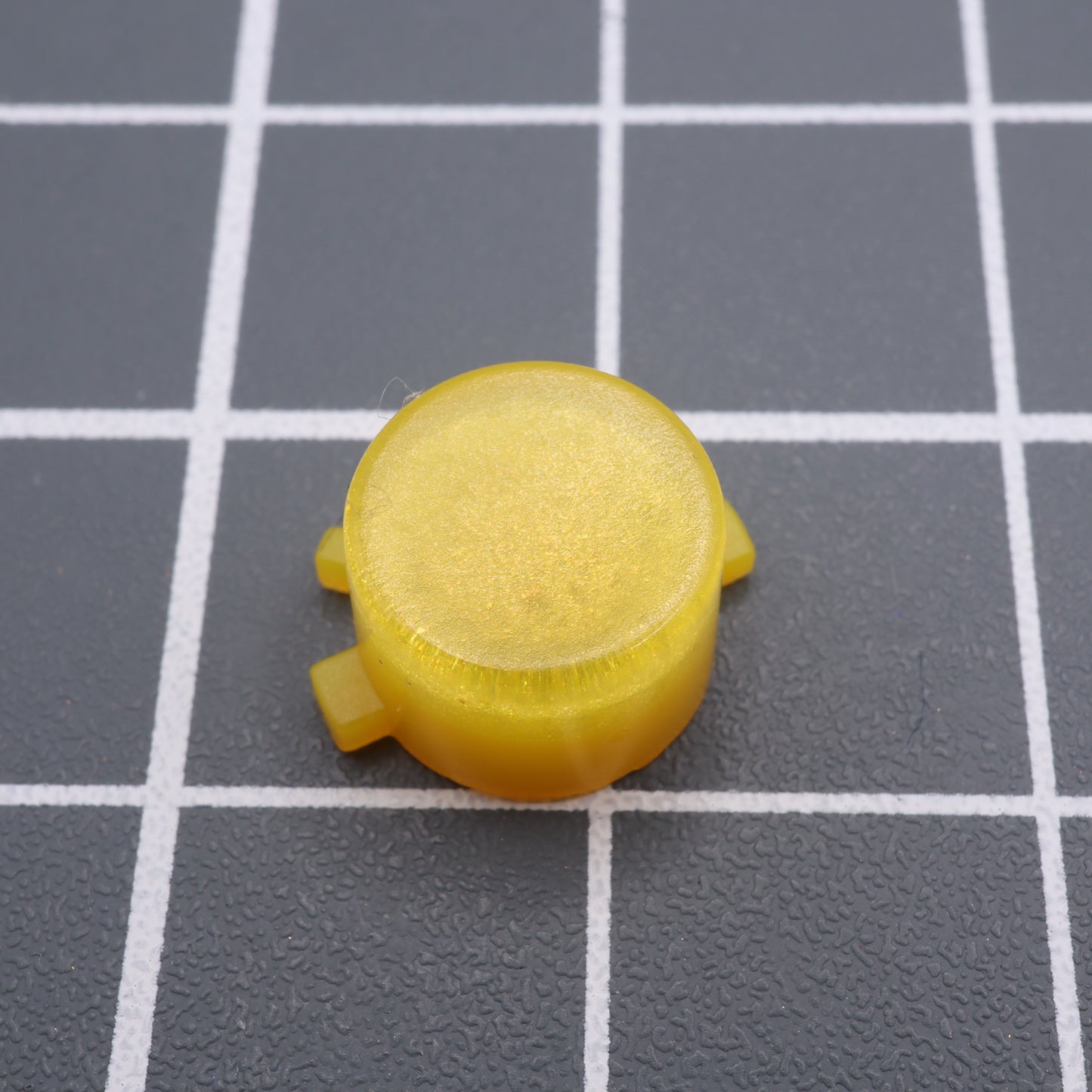 The Analogue Pocket - Custom Button in Lemon Candy is a round yellow plastic button with a flat top and two side tabs, set against a gray grid background, showcasing an intricately handmade design akin to crafting custom-cast buttons.