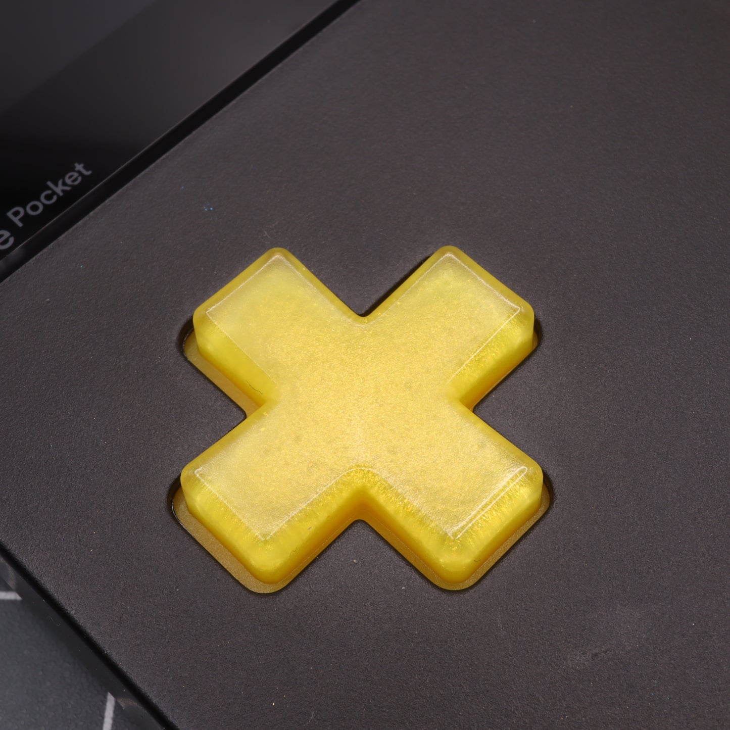 Close-up of a Lemon Candy custom button, translucent and cross-shaped, on the Analogue Pocket's sleek black surface, crafted using handmade molds.