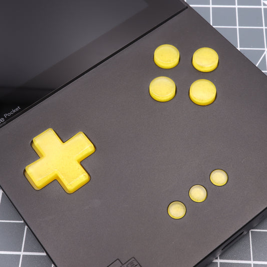 A close-up of the Analogue Pocket - Custom Button - Lemon Candy, a handheld gaming device with a black casing and custom-cast yellow buttons, including a directional pad and circular action buttons, resting on a grid-patterned surface.