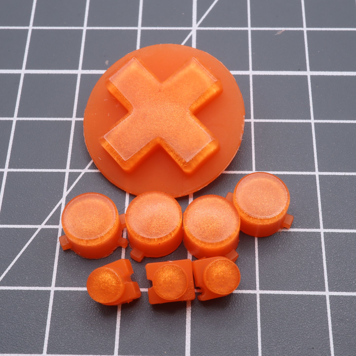 Orange Candy custom-cast buttons from the Analogue Pocket, featuring both button and D-pad designs, are set against a grid surface.