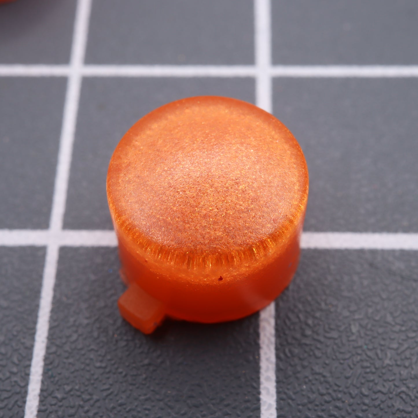 Close-up of an Orange Candy custom-cast button for the Analogue Pocket on a gray grid-patterned surface.