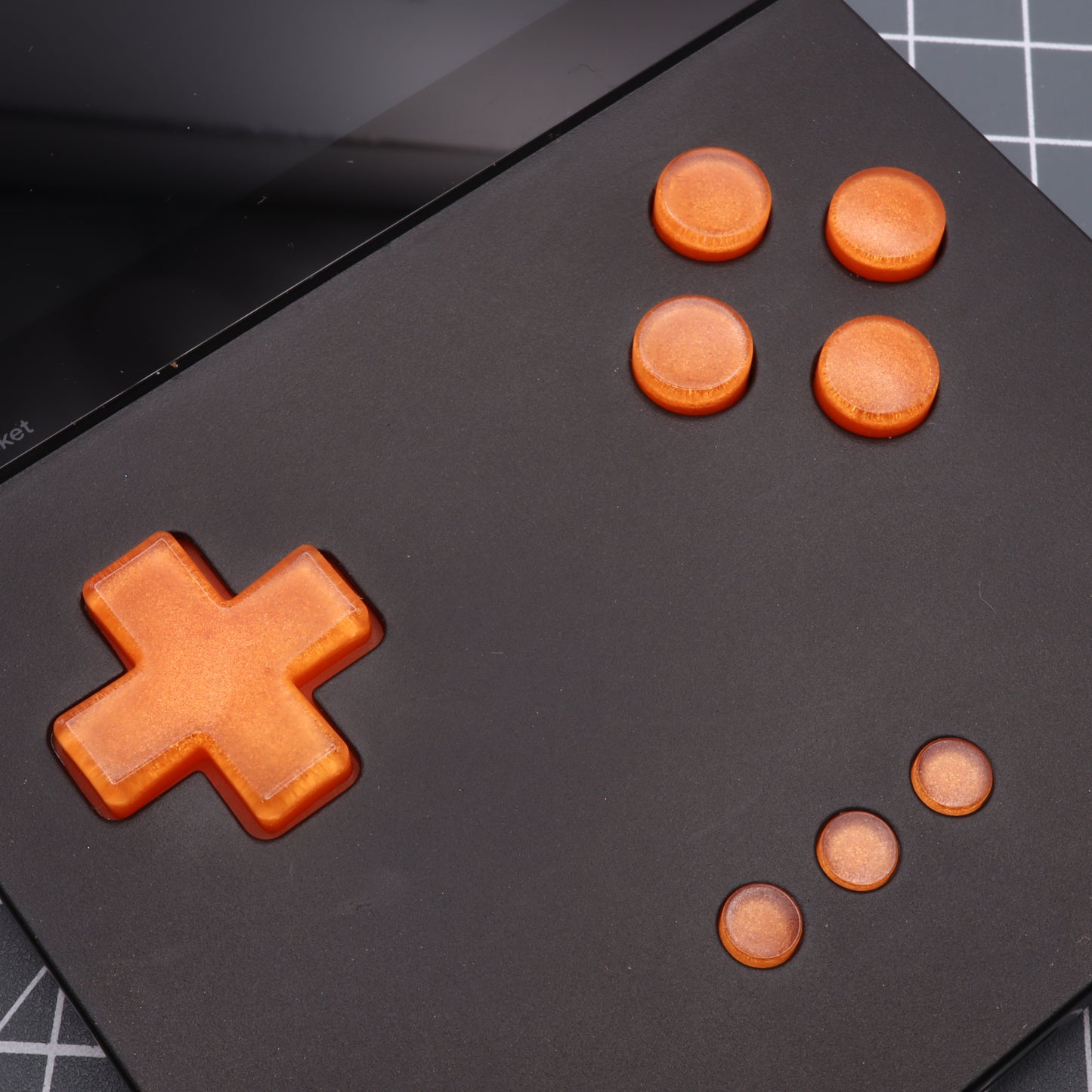 Close-up of a black gaming device with striking orange Analogue Pocket - Custom Button - Orange Candy buttons in a cross and circular pattern on a grid background.