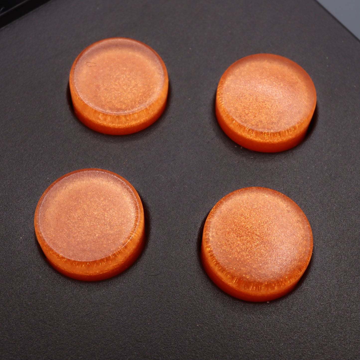 The Analogue Pocket Custom Buttons in Orange Candy, four in total, shimmer with a glossy finish while displayed on a dark surface.