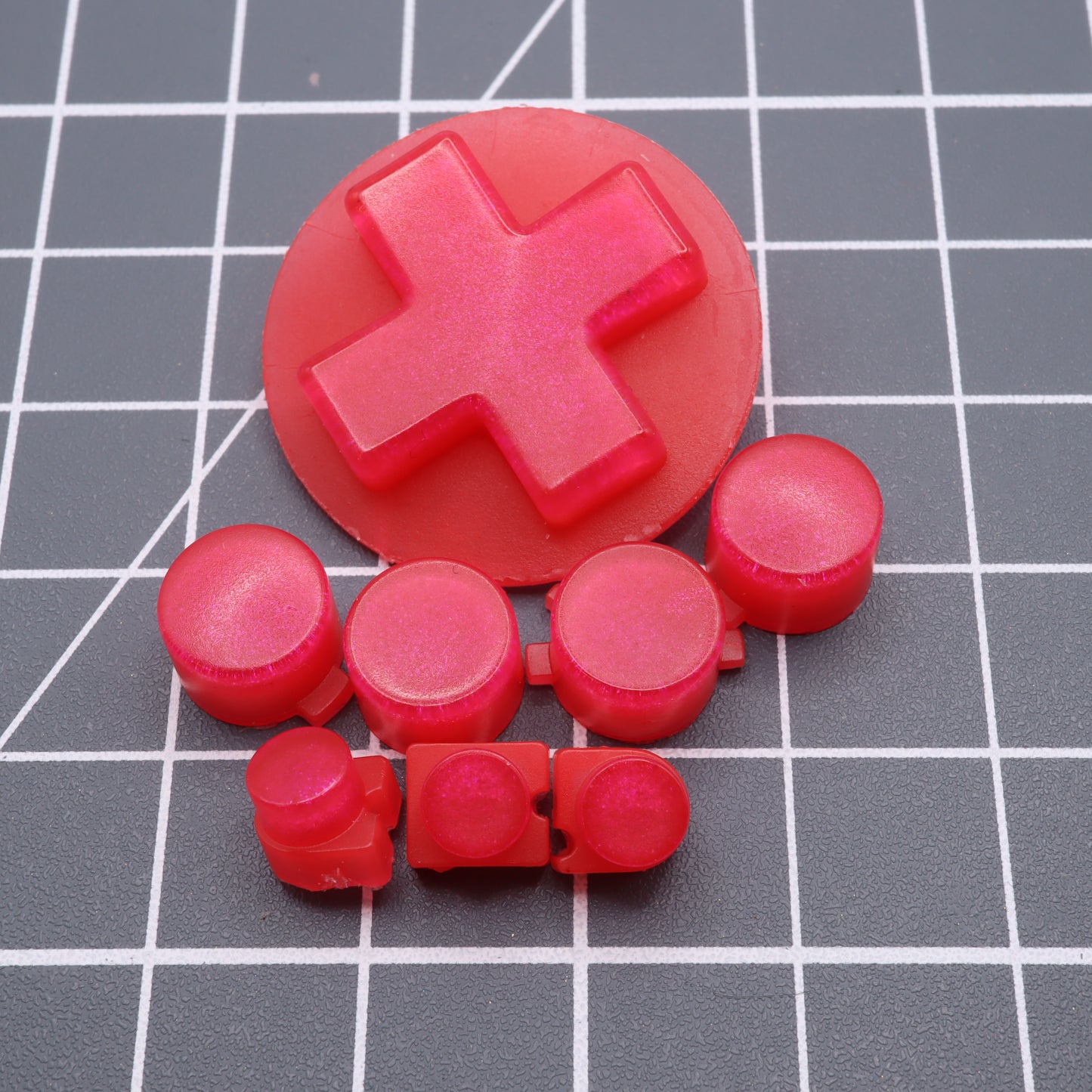 Red, round, plus-shaped Raspberry Candy buttons crafted from custom molds are set on a grid-patterned surface, reminiscent of Analogue Pocket's design.