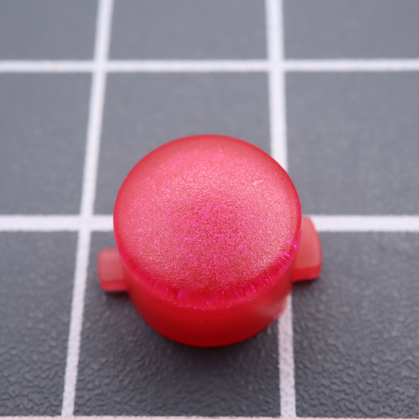 A Raspberry Candy custom button from Analogue Pocket, featuring a red plastic cap with a textured surface, sits on a gray grid background.