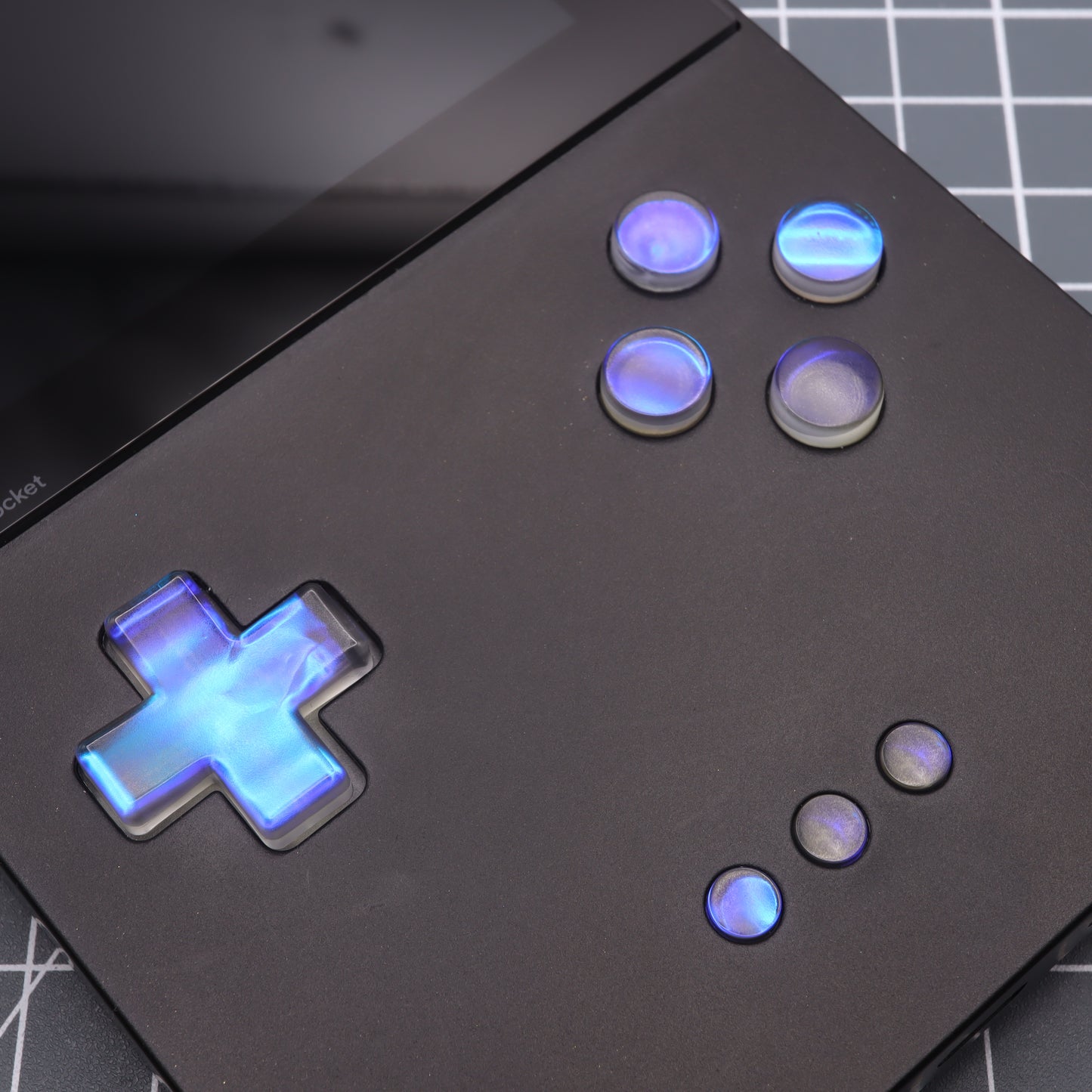 Close-up of the Analogue Pocket - Custom Button - Midnight Opal, a black handheld gaming device featuring glowing blue buttons in a D-pad layout and circular buttons against a grid background.