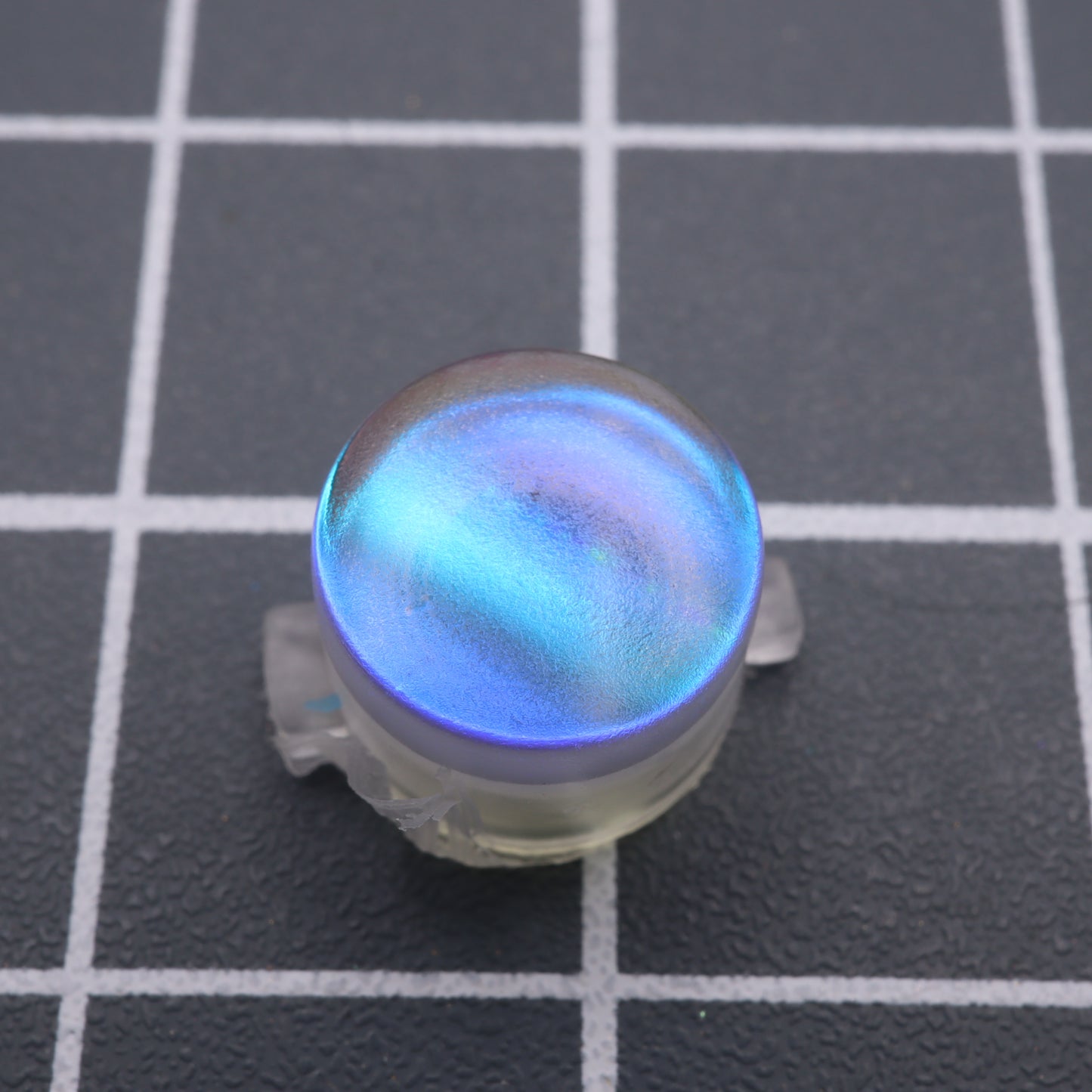 A round, iridescent blue object crafted from handmade molds, known as the Analogue Pocket - Custom Button - Midnight Opal, rests elegantly on a black grid-patterned surface.