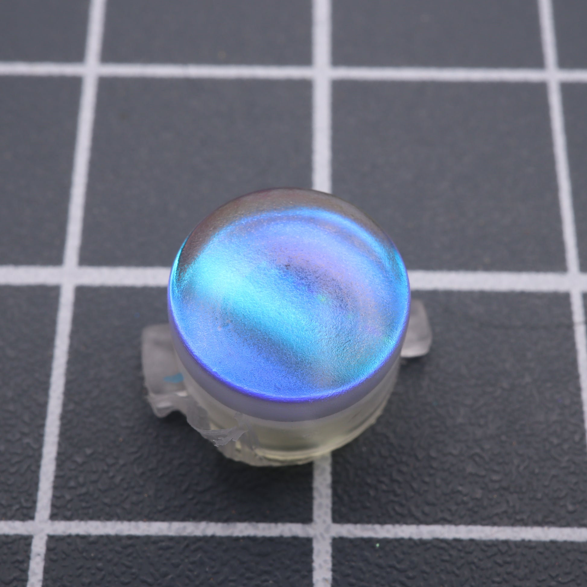 A round, iridescent blue object crafted from handmade molds, known as the Analogue Pocket - Custom Button - Midnight Opal, rests elegantly on a black grid-patterned surface.