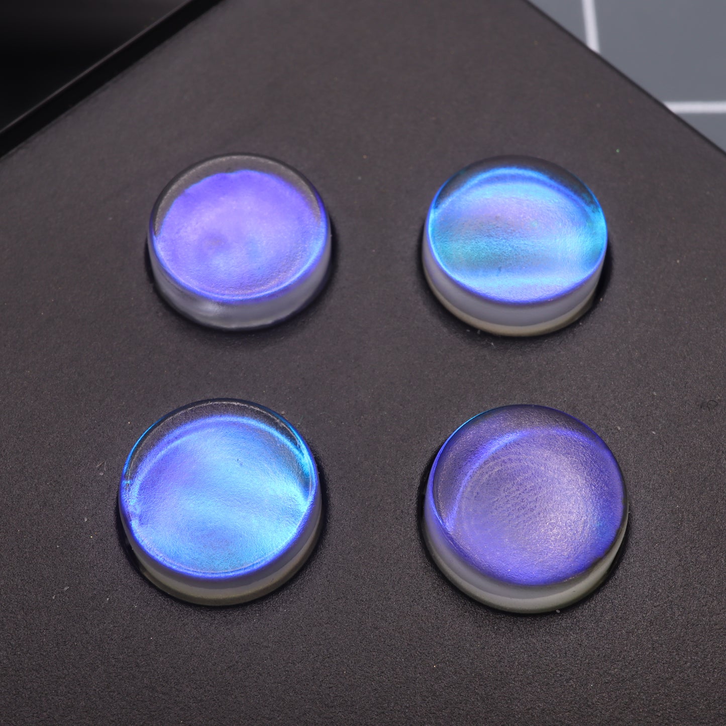 Four Midnight Opal Analogue Pocket custom buttons, iridescent blue and circular with exquisite detail, rest on a dark surface.
