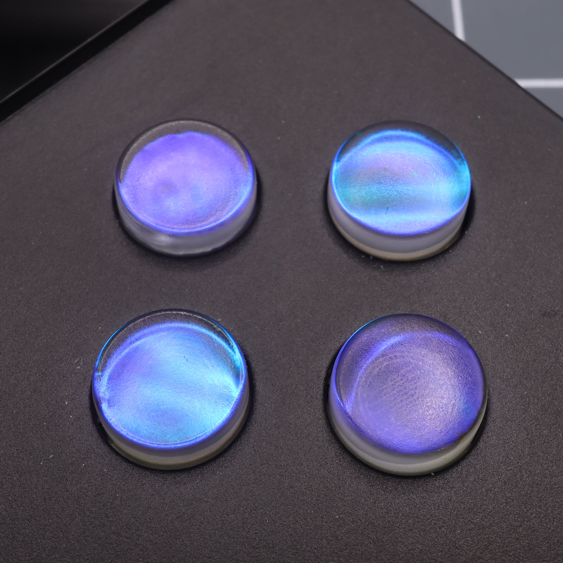 Four Midnight Opal Analogue Pocket custom buttons, iridescent blue and circular with exquisite detail, rest on a dark surface.