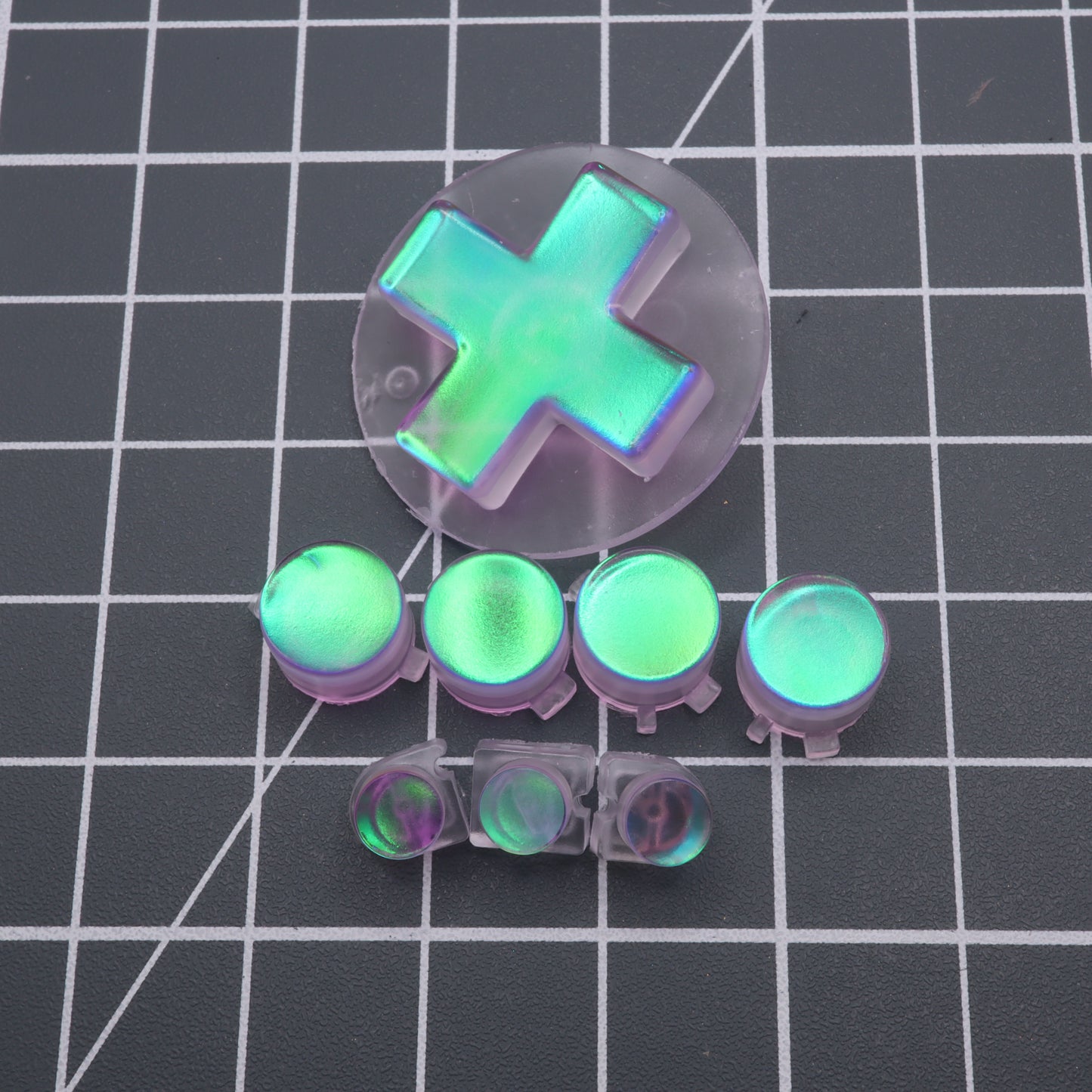 The Cool Opal custom-cast buttons and D-pad for the Analogue Pocket gaming controller shimmer iridescently on a grid-patterned surface.