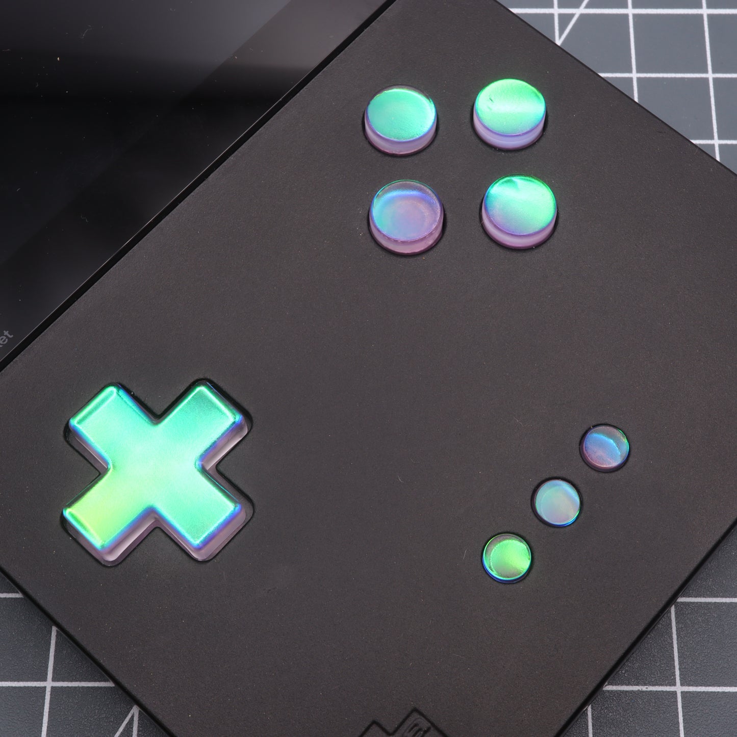 A close-up of the Analogue Pocket - Custom Button in Cool Opal, a black handheld gaming device with iridescent buttons and directional pad, placed on a grid-patterned surface.