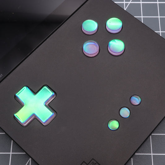 A close-up of the Analogue Pocket - Custom Button in Cool Opal, a black handheld gaming device with iridescent buttons and directional pad, placed on a grid-patterned surface.