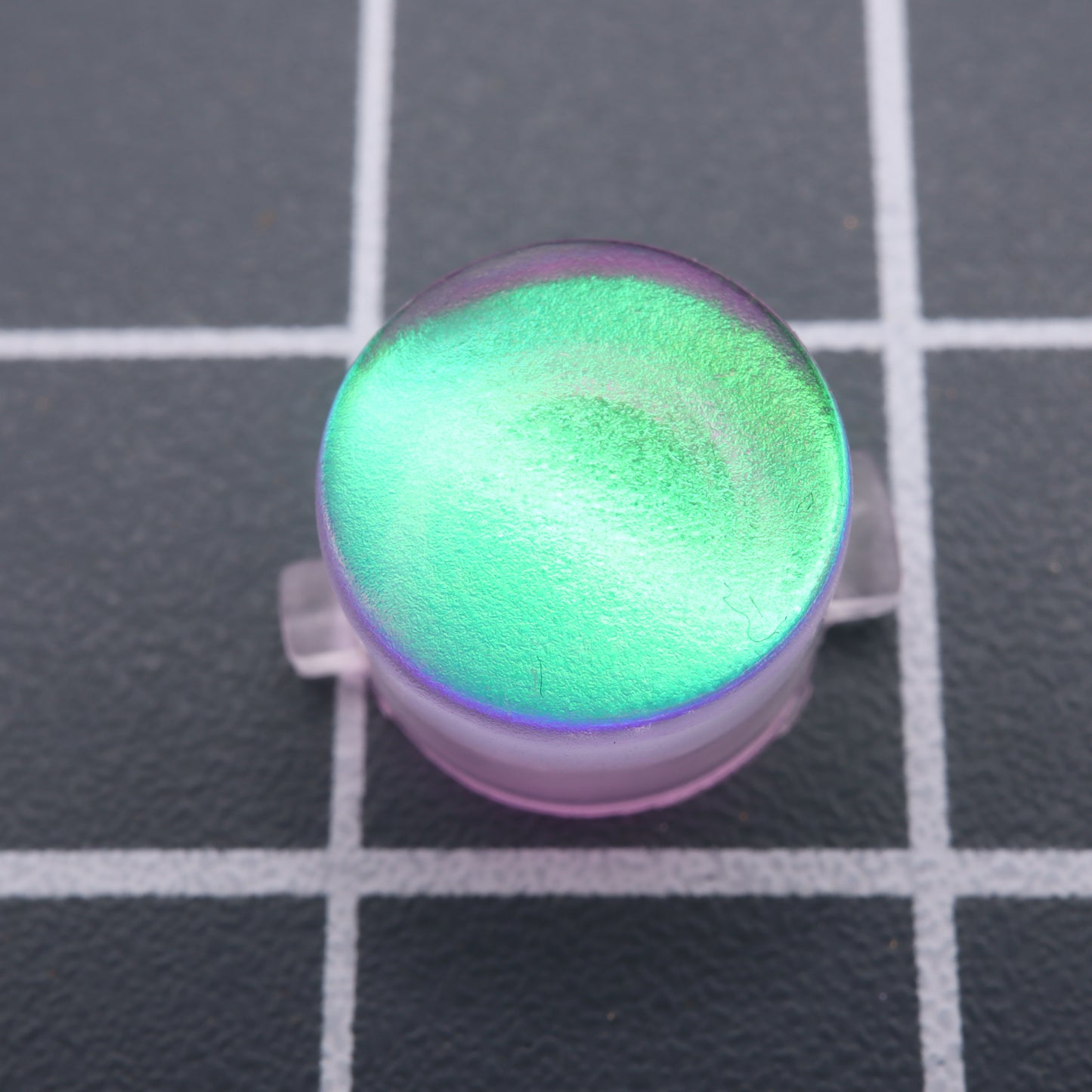 A round, iridescent green object called the Analogue Pocket - Custom Button in Cool Opal gleams with a reflective surface on a grid-patterned background.