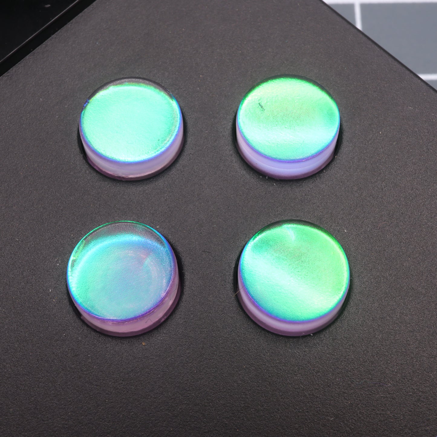 Four round, iridescent green and purple buttons, resembling the Cool Opal custom buttons for the Analogue Pocket, are placed on a black surface.