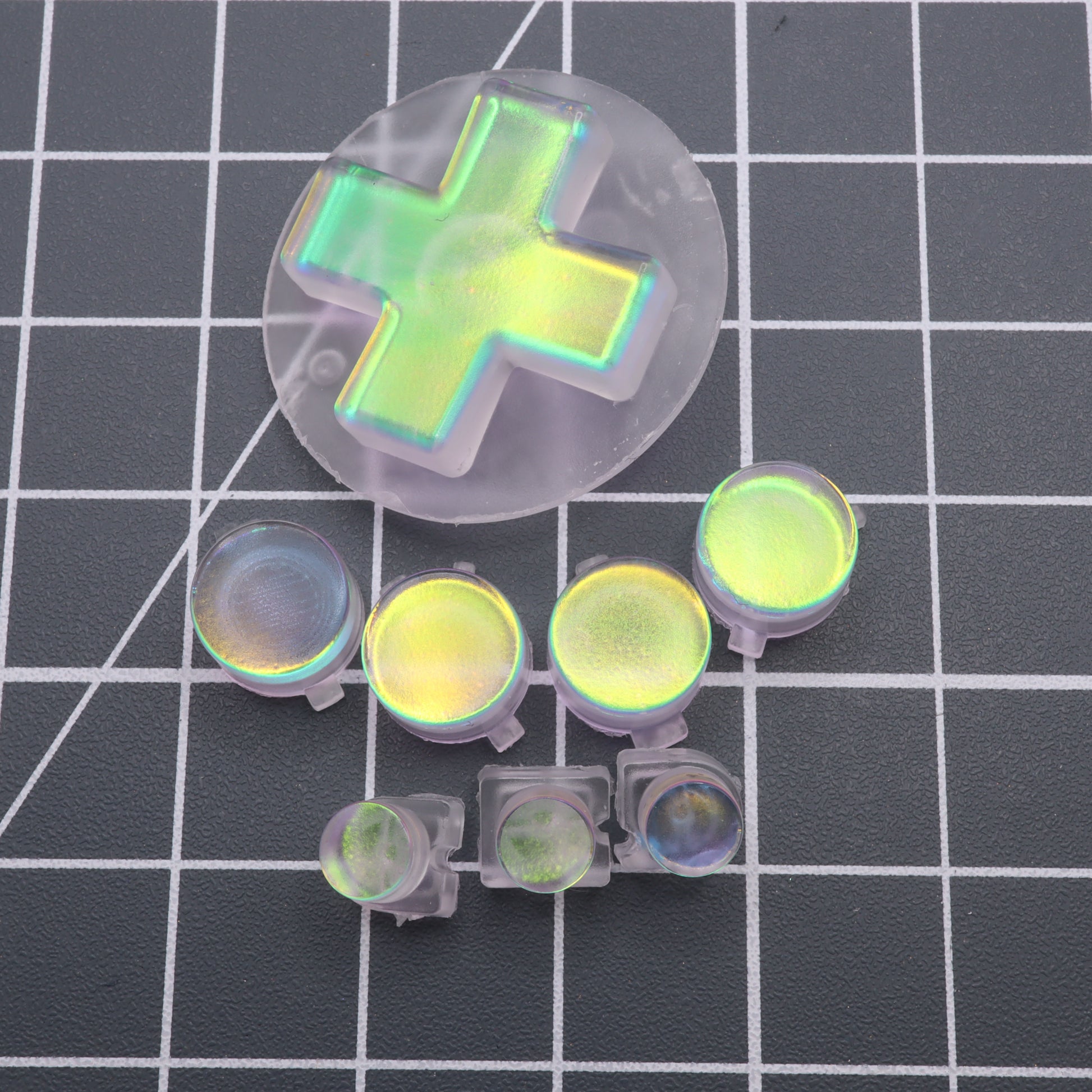 A set of Sunset Opal Analogue Pocket custom buttons, made from iridescent, translucent resin in diverse shapes and sizes, neatly displayed on a grid-patterned surface.