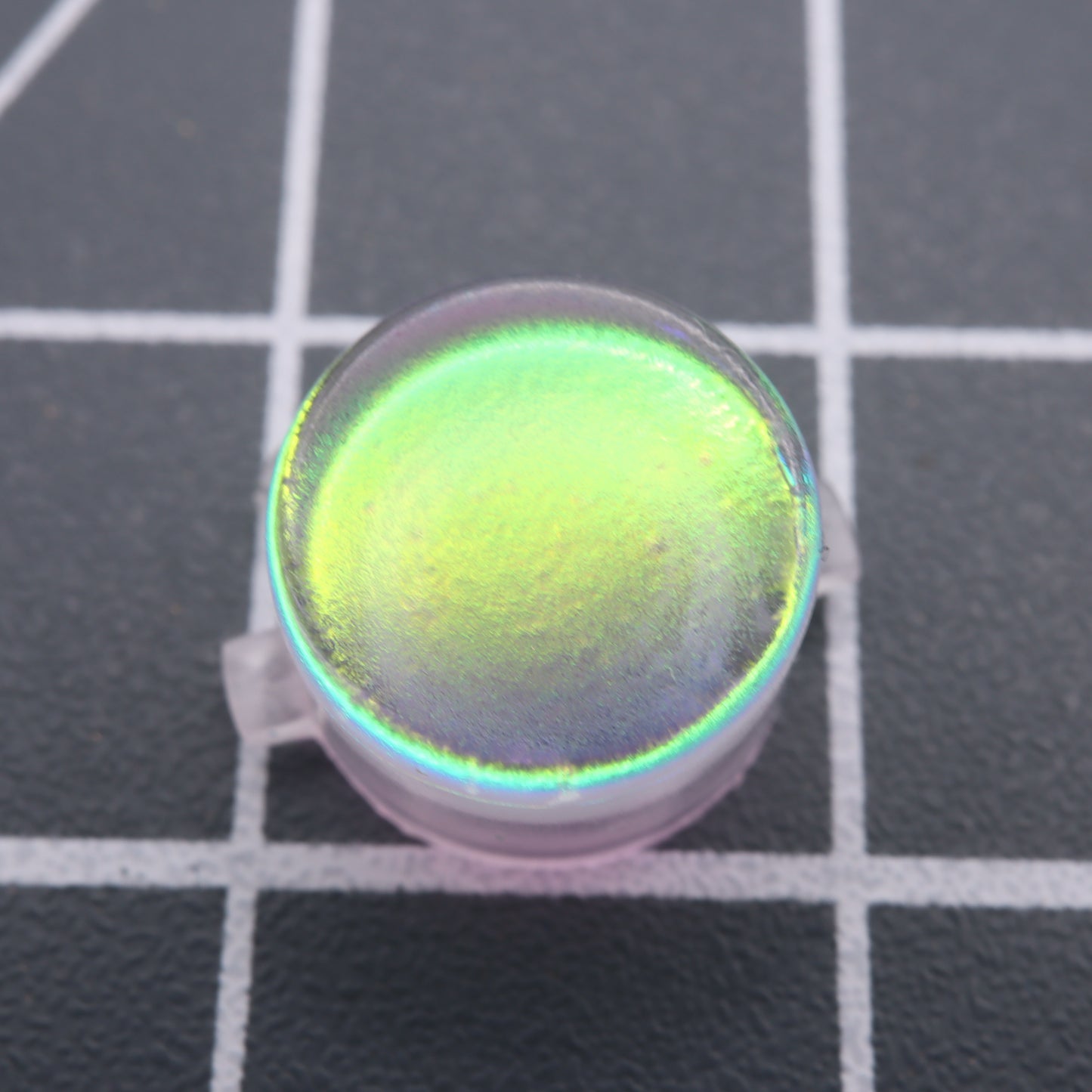 A close-up of the Analogue Pocket - Custom Button - Sunset Opal on a grid-patterned surface, showcasing a mesmerizing green and yellow gradient.