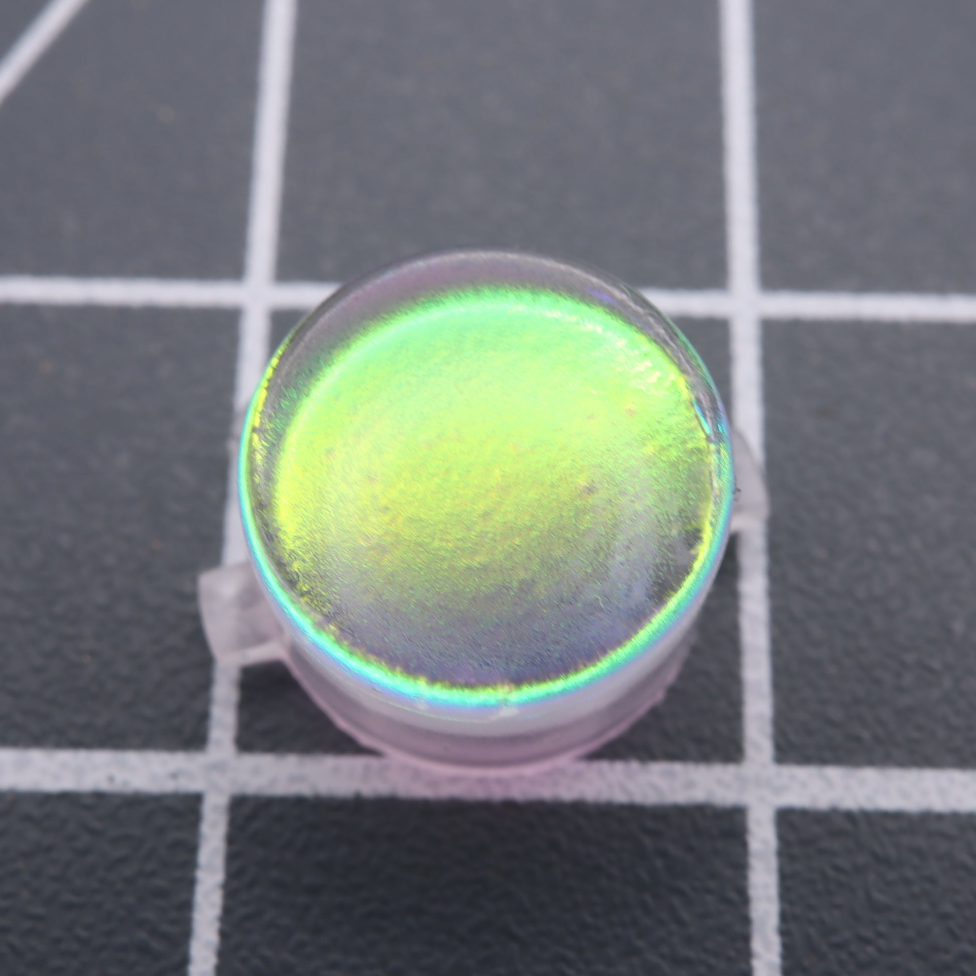 A close-up of the Analogue Pocket - Custom Button - Sunset Opal on a grid-patterned surface, showcasing a mesmerizing green and yellow gradient.
