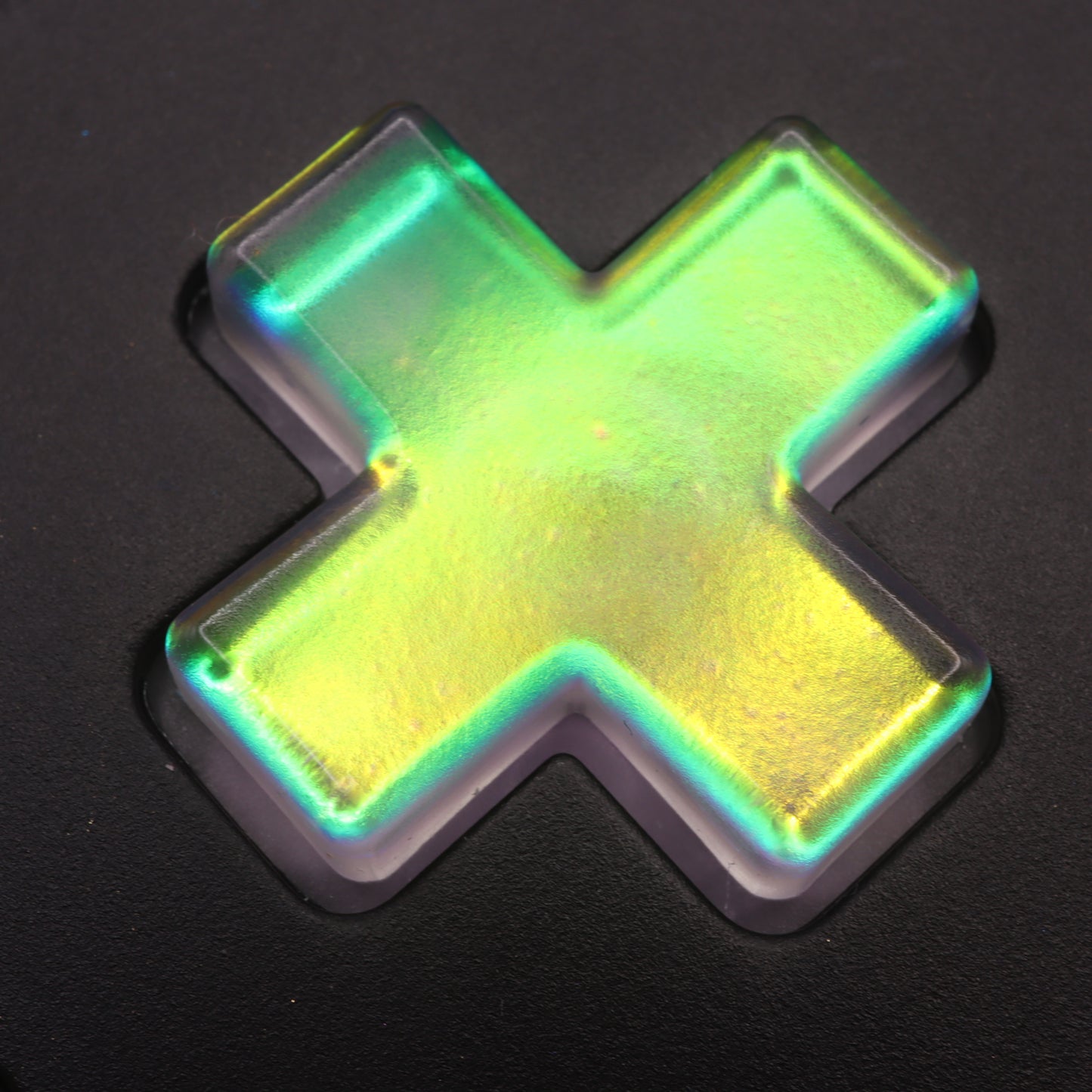 A glowing, iridescent green and yellow X-shaped object similar to the Sunset Opal custom-cast resin button from Analogue Pocket, set against a dark background.