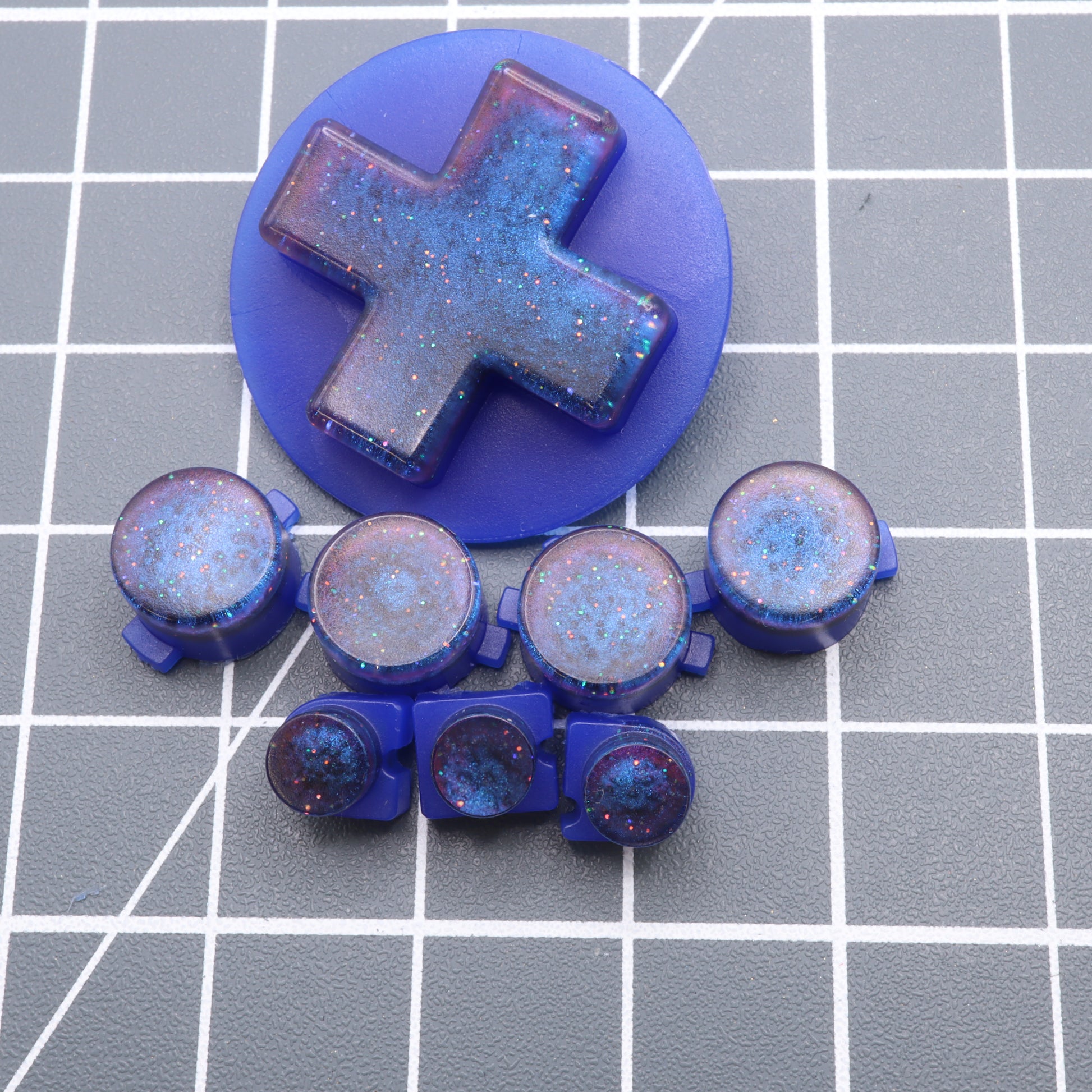 The "Cosmic Blue" buttons for the Analogue Pocket are custom-cast from blue resin with glitter, beautifully presented on a grid background, reflecting craftsmanship from handmade molds.