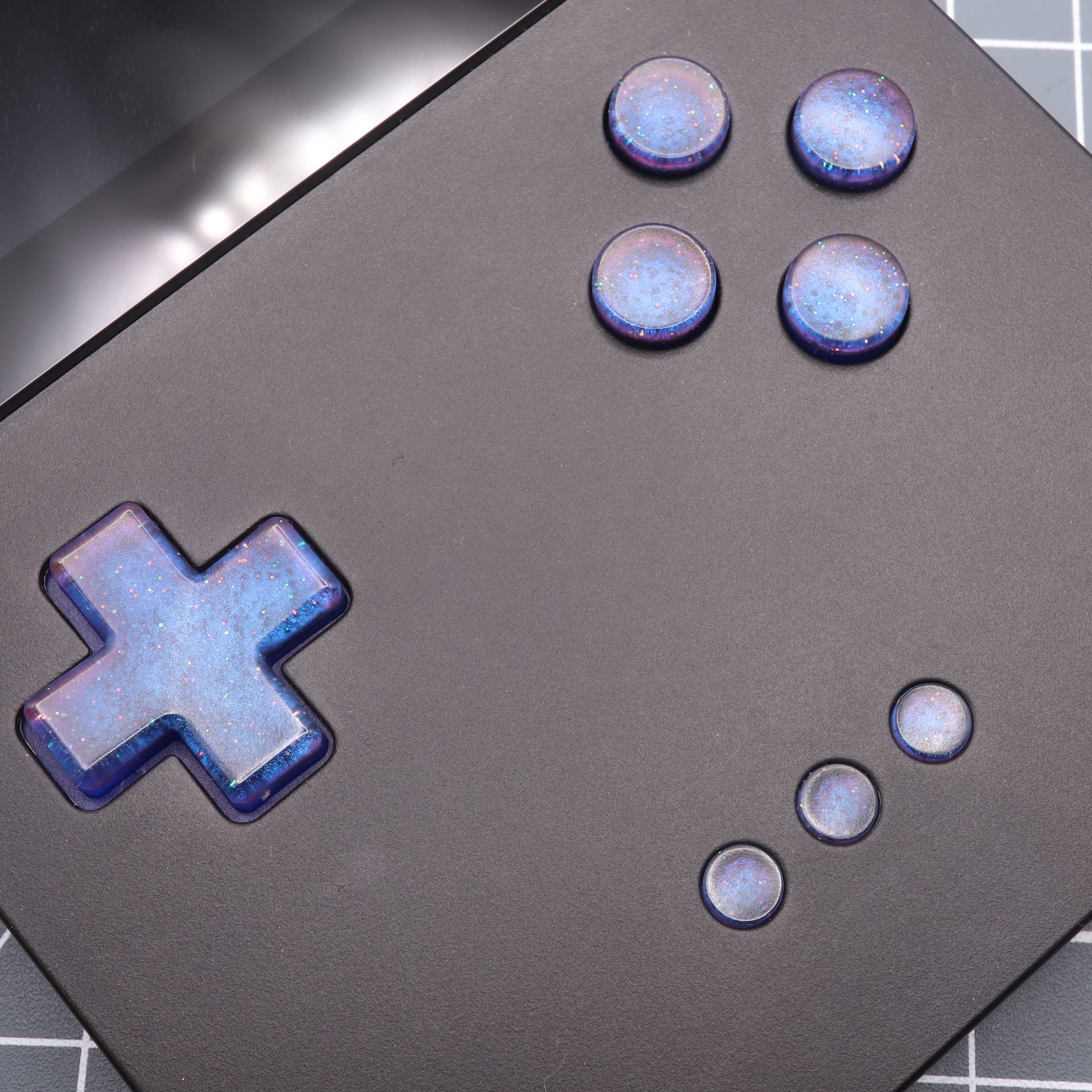 The Analogue Pocket - Custom Button - Cosmic Blue features a black gaming device with custom-cast buttons, a glossy D-pad, and four round blue galaxy-patterned buttons.