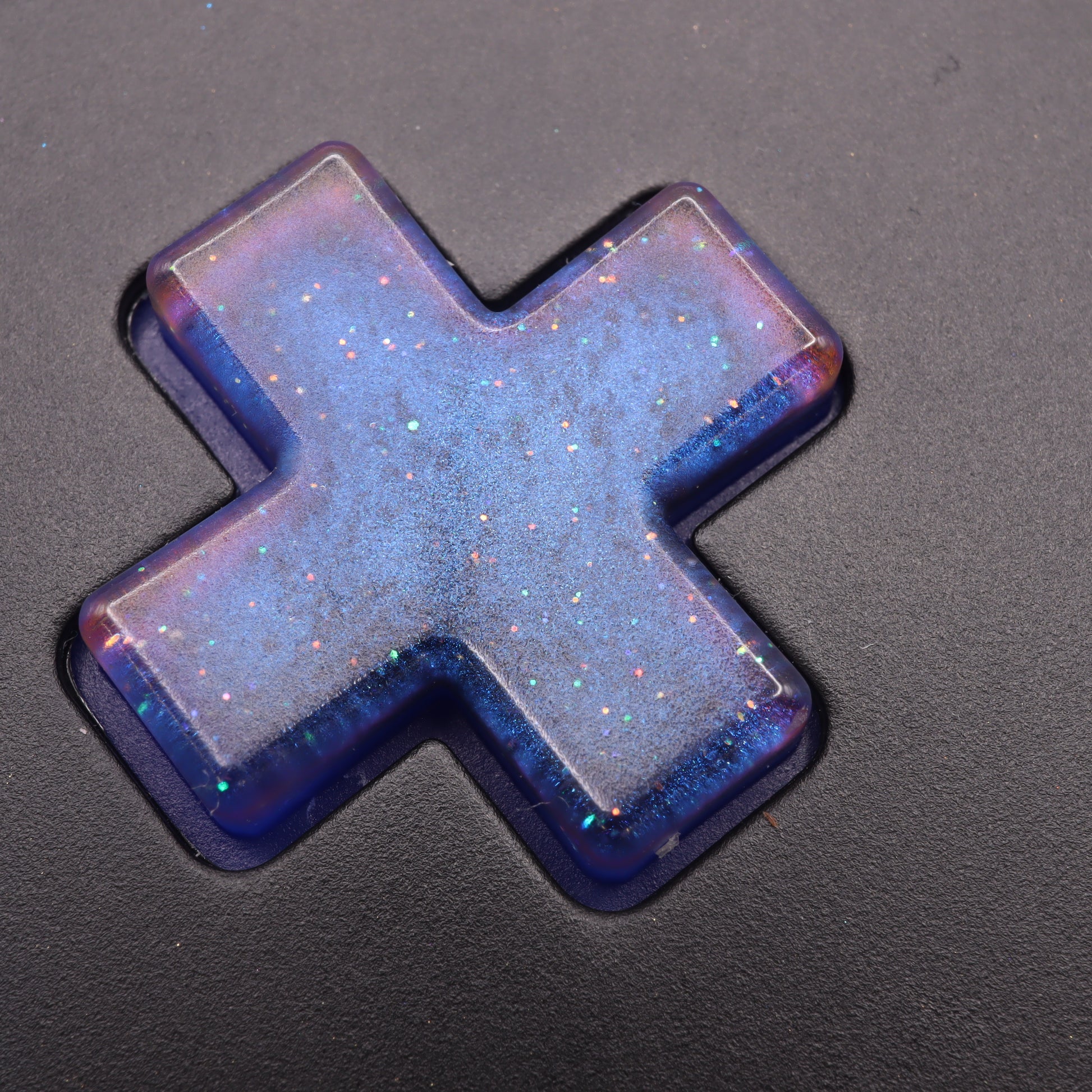 The Analogue Pocket - Custom Button - Cosmic Blue is a translucent, galaxy-themed plus-shaped keycap created with handmade molds in blue and purple hues. It features embedded glitter that shimmers beautifully on a black surface.