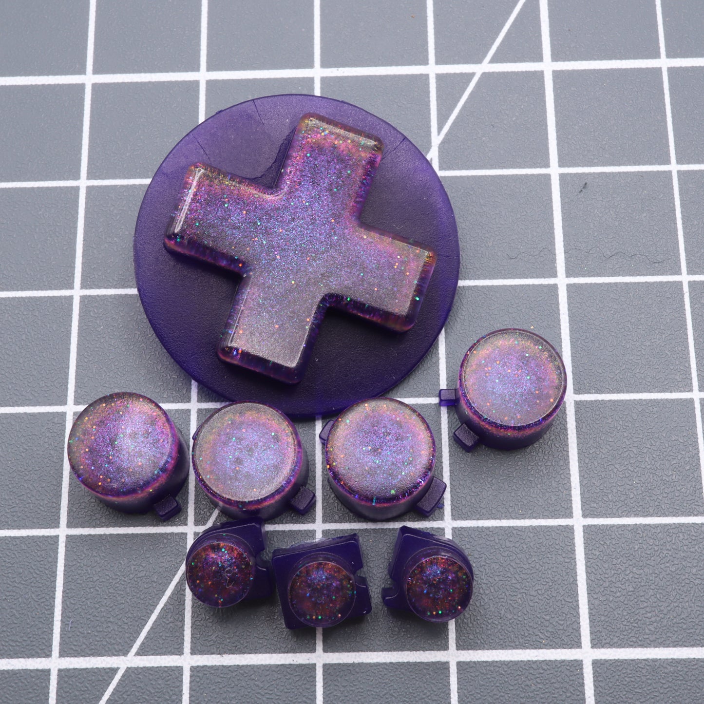 The Cosmic Purple custom-cast buttons for Analogue Pocket, including a D-pad and several round ones, are displayed on a gray grid surface, highlighting exquisite craftsmanship perfect for any setup.