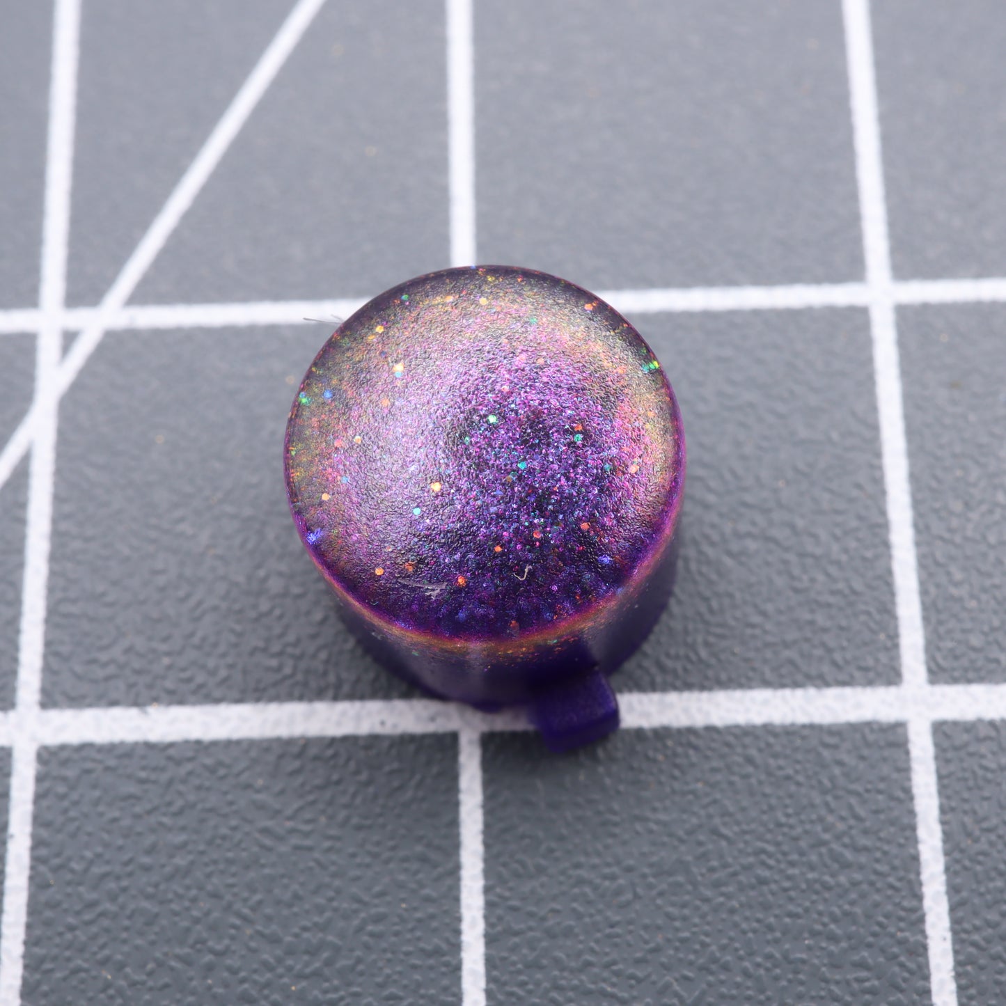 A Cosmic Purple Analogue Pocket custom button, round and glittery, rests elegantly on a gray grid-patterned background.
