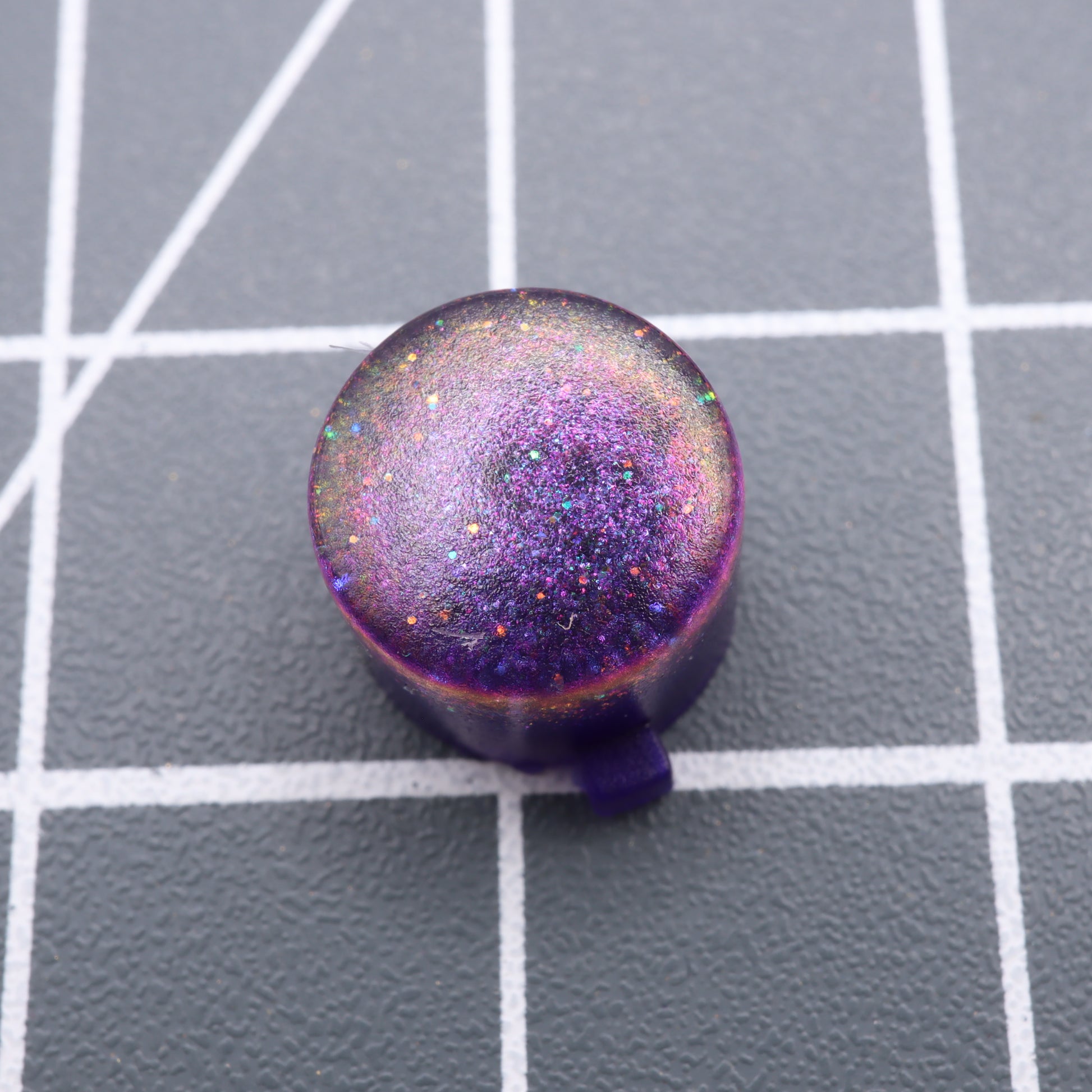 A Cosmic Purple Analogue Pocket custom button, round and glittery, rests elegantly on a gray grid-patterned background.