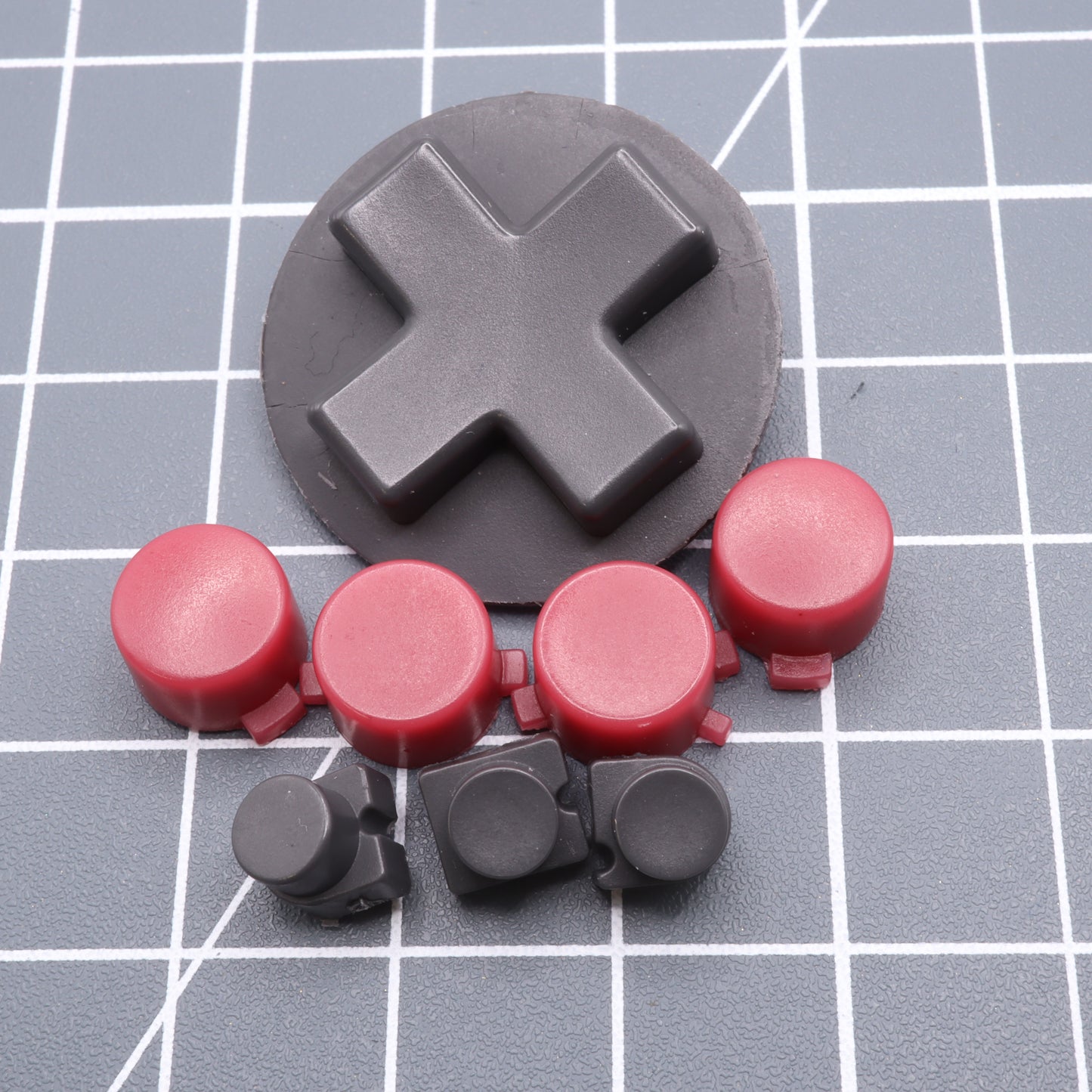 A large black cross-shaped key and smaller red and black circular keys rest on a grid-patterned surface, resembling custom-cast Analogue Pocket - Custom Button - Game Boy DMG Style buttons.