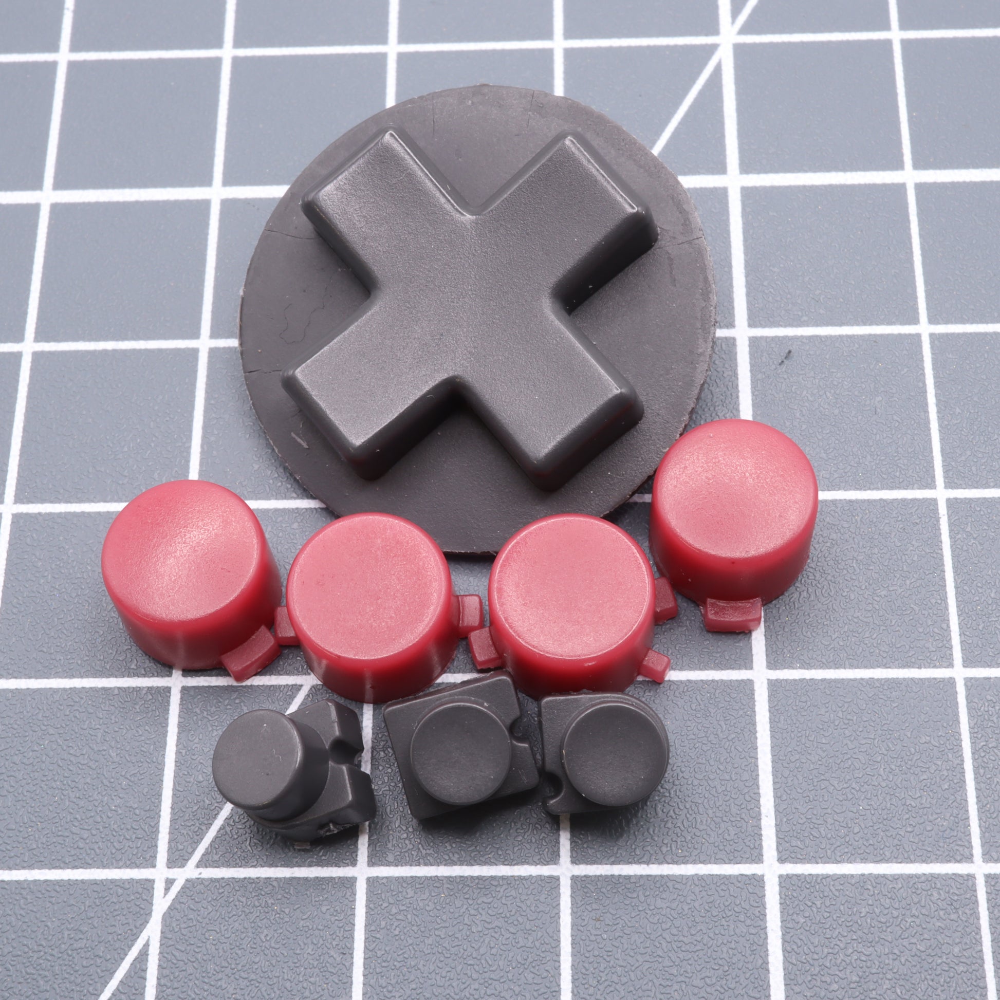 A large black cross-shaped key and smaller red and black circular keys rest on a grid-patterned surface, resembling custom-cast Analogue Pocket - Custom Button - Game Boy DMG Style buttons.