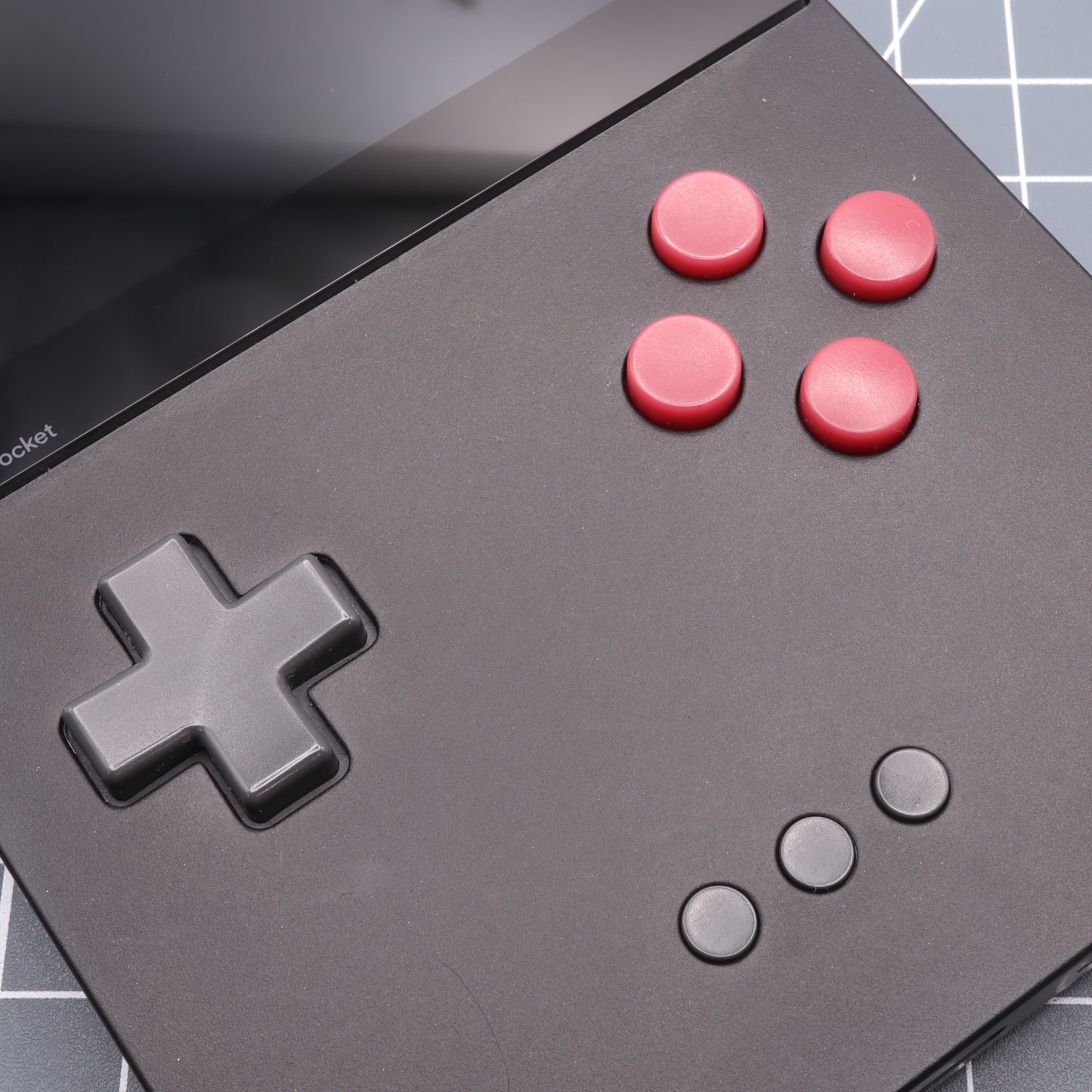 Close-up of the Analogue Pocket - Custom Button - Game Boy DMG Style, a black gaming handheld device featuring a cross-shaped directional pad on the left and four custom-cast red buttons on the right, with three small buttons below.
