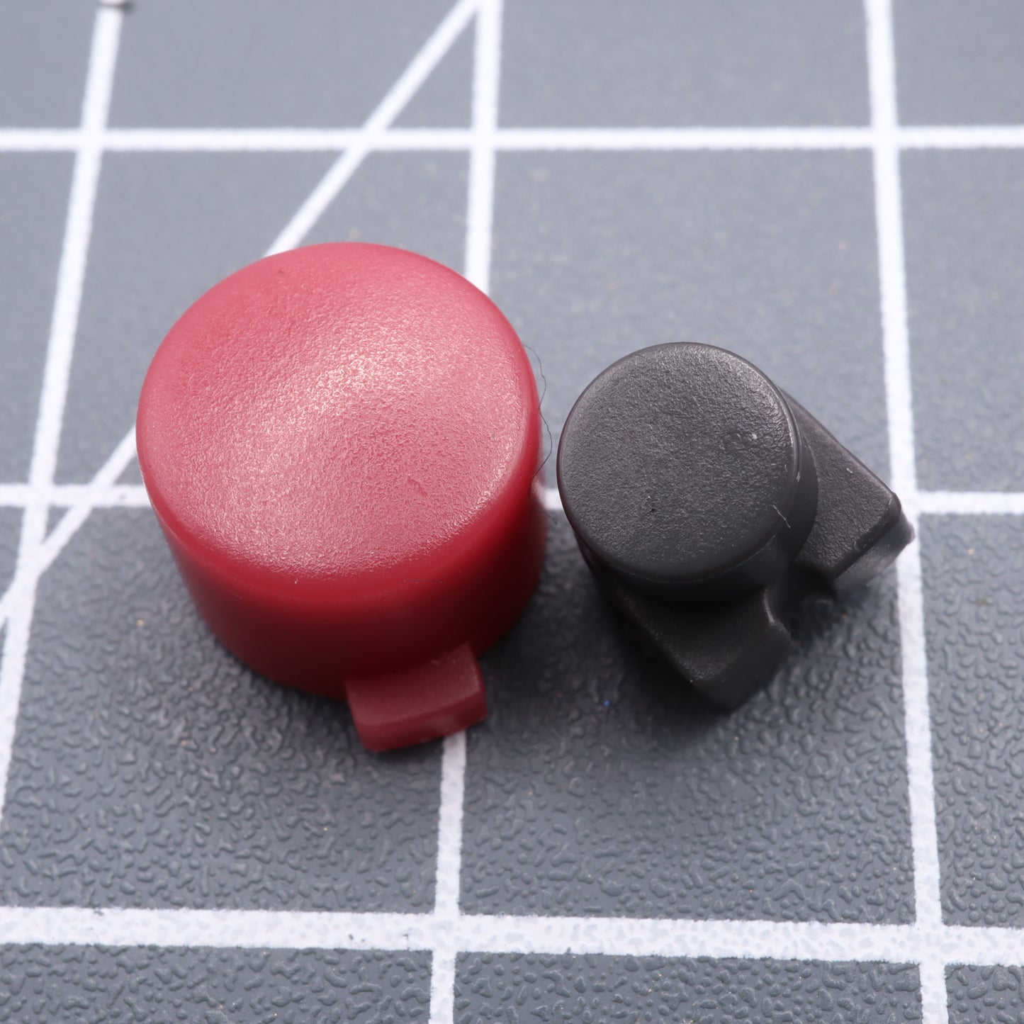 A red circular keycap and a black keycap with a notch, crafted from custom-cast buttons of the Analogue Pocket - Custom Button - Game Boy DMG Style, rest on a grid-patterned surface.