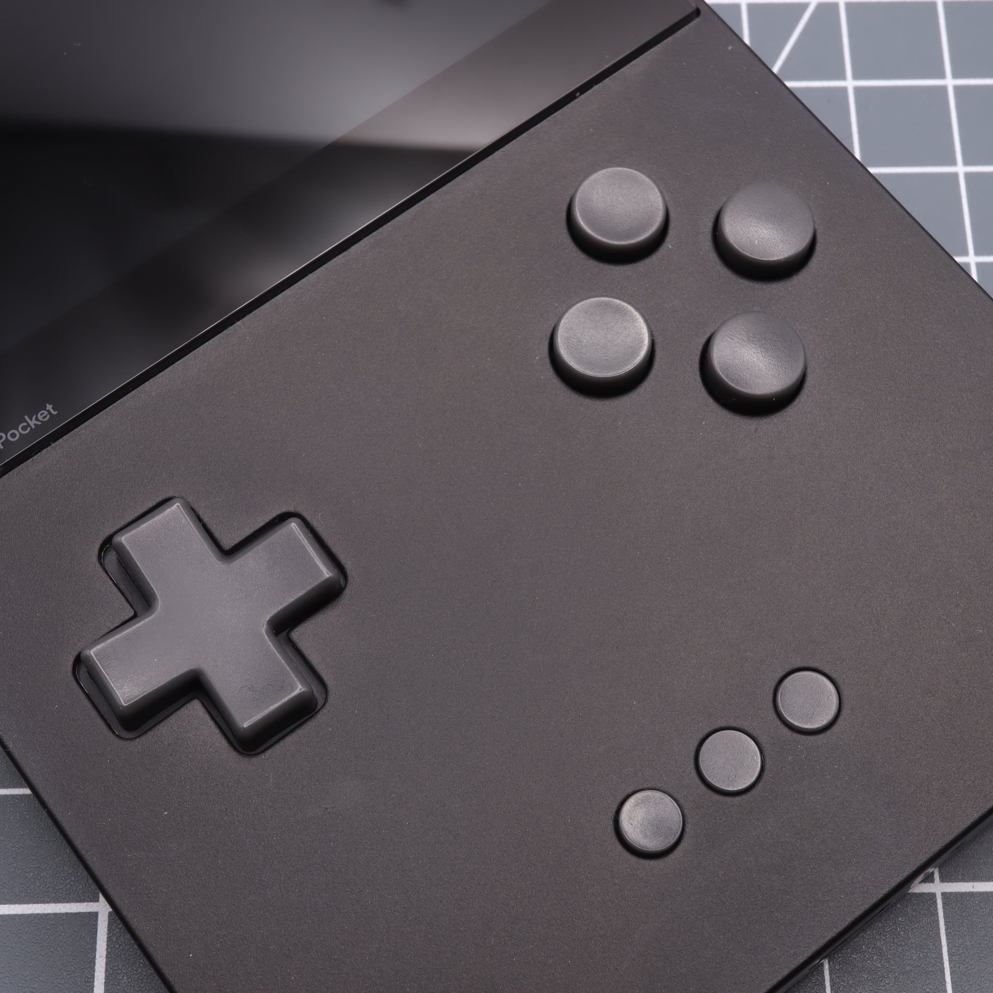 Close-up of the Analogue Pocket - Custom Button - DMG Play it Loud Style, a black handheld gaming device with resin buttons, a screen, directional pad, and six circular buttons on a grid-patterned background.