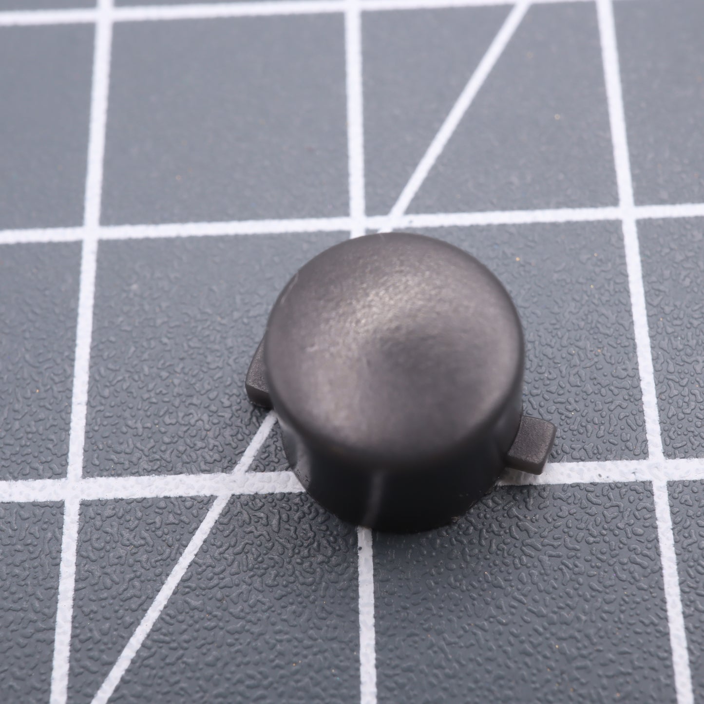 A DMG Play it Loud Style custom black round resin button with side tabs from an Analogue Pocket is on a gray grid surface.